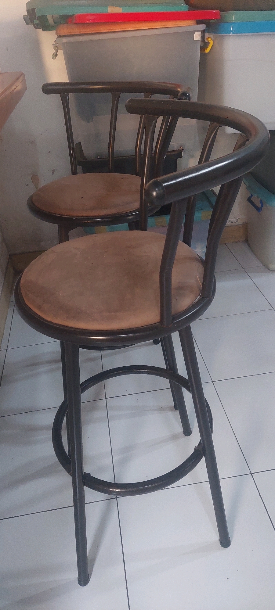 The image displays two dark brown bar stools with round, light brown upholstered seats. The stools have curved backs and metal legs with a circular footrest. They are placed on a tiled floor. In the background, there are stacked plastic containers with lids in various colors, including green, red, and blue. A white wall is visible behind the containers. The overall setting appears to be an indoor storage area or a room with utilitarian furniture. The specific location in Lumajang, Indonesia, is not discernible from the visual information provided in the image.