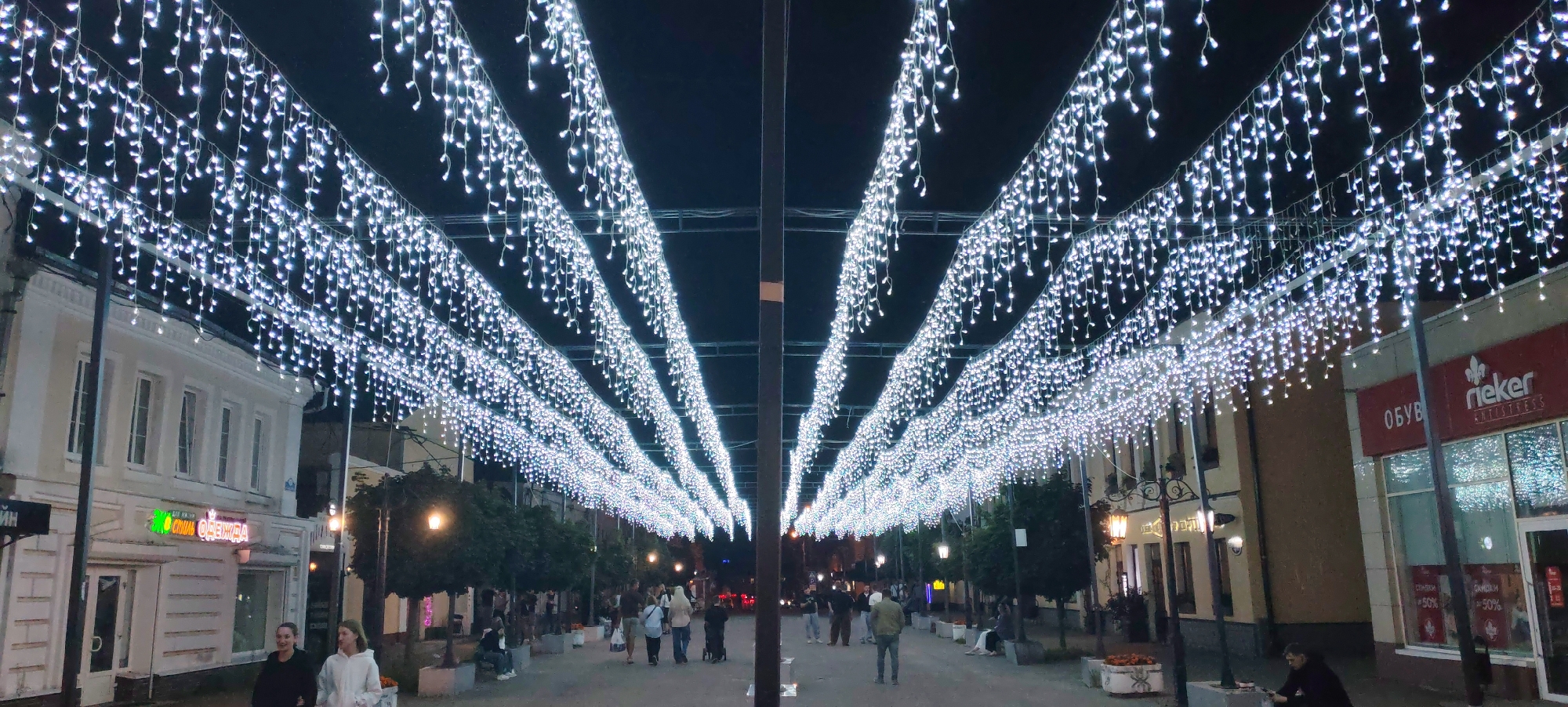 The image depicts a street scene at night in Kaluga, Russia, adorned with numerous strings of white icicle lights creating a luminous canopy over a pedestrian walkway. Buildings with traditional architecture line both sides of the street, some featuring shopfronts with illuminated signs. People are seen walking along the street, some in groups, others individually. The lighting from the icicle strings casts a bright glow, illuminating the walkway and creating a festive atmosphere. Trees are planted along the sides of the walkway, adding a touch of nature to the urban setting. The overall scene suggests a well-lit and vibrant public space, possibly a shopping or entertainment district, enhanced by decorative lighting.