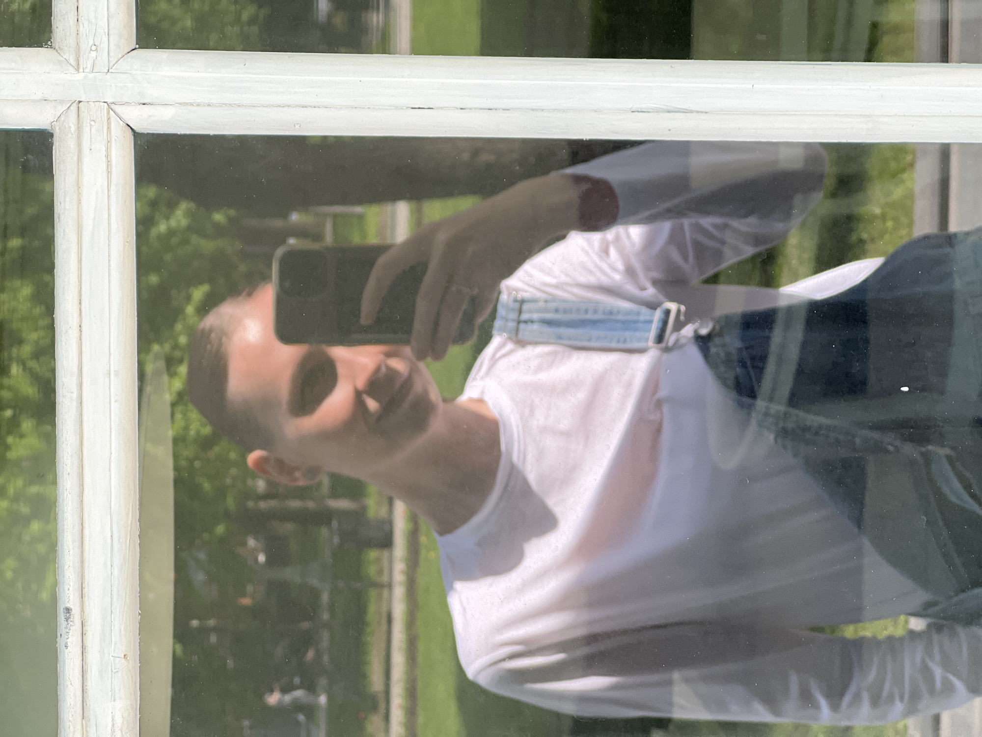 A reflection in a window pane shows a person holding a smartphone to take a picture of themselves. The person is wearing a white long-sleeved shirt and dark overalls with light blue straps. The background visible through the window includes greenery, suggesting a park or garden. The white frame of the window is visible. I cannot discern a specific location in Moscow, Russia from this image alone.