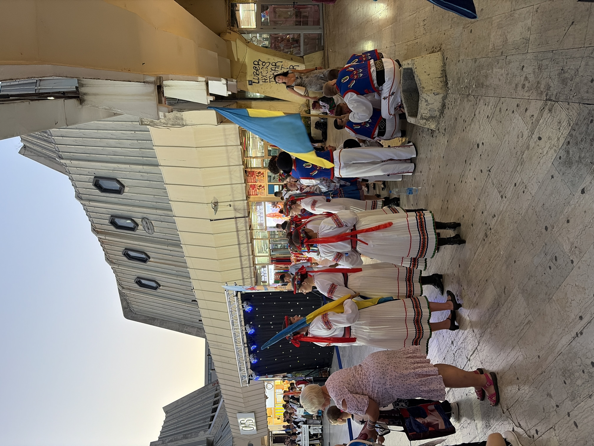 In the city of Bar, Montenegro, an outdoor gathering takes place beneath a modernist architectural structure featuring corrugated metal and angular concrete elements. The scene is dominated by a group of people, many of whom are dressed in traditional Ukrainian attire. Several individuals, primarily women, are wearing white embroidered dresses with red accents and headbands. A group of young men and women are also present, with some holding a Ukrainian flag, a blue and yellow bicolor. They appear to be part of a cultural performance or celebration. In the background, a stage with a black backdrop and blue lights is visible, suggesting an event is or will be taking place. The ground is paved with large, light-colored stone tiles, and there is a small, stone basin-like structure in the foreground. The lighting suggests it is late afternoon or early evening. Shops and entrances to buildings are visible in the background, indicating an urban commercial area.