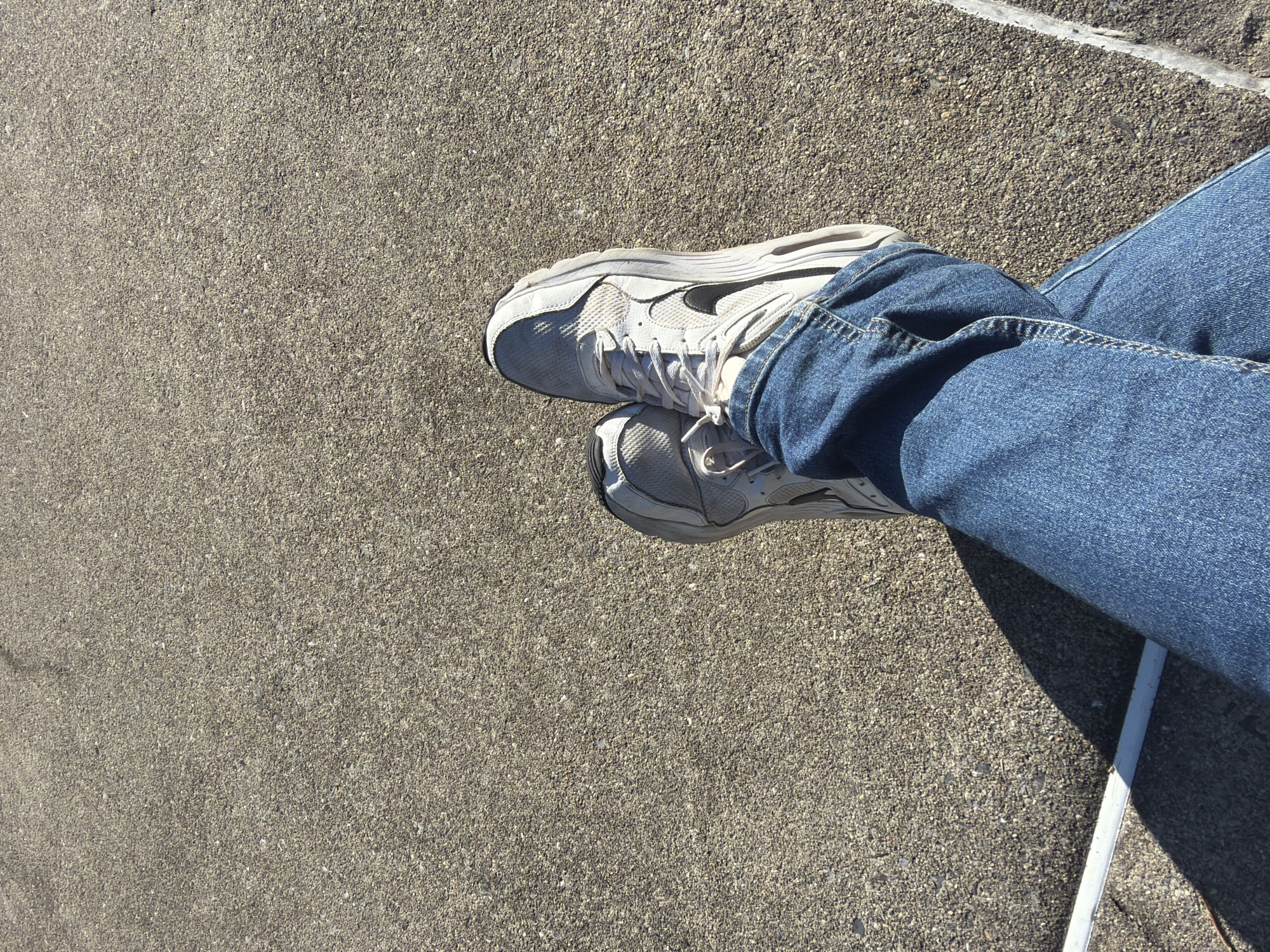 The image shows a person sitting with legs crossed, wearing blue jeans and white athletic shoes with a black swoosh logo, on a concrete surface. The setting appears to be outdoors, with a sidewalk or paved area visible. A white object or edge is seen near the legs. The location is Edison, United States of America.