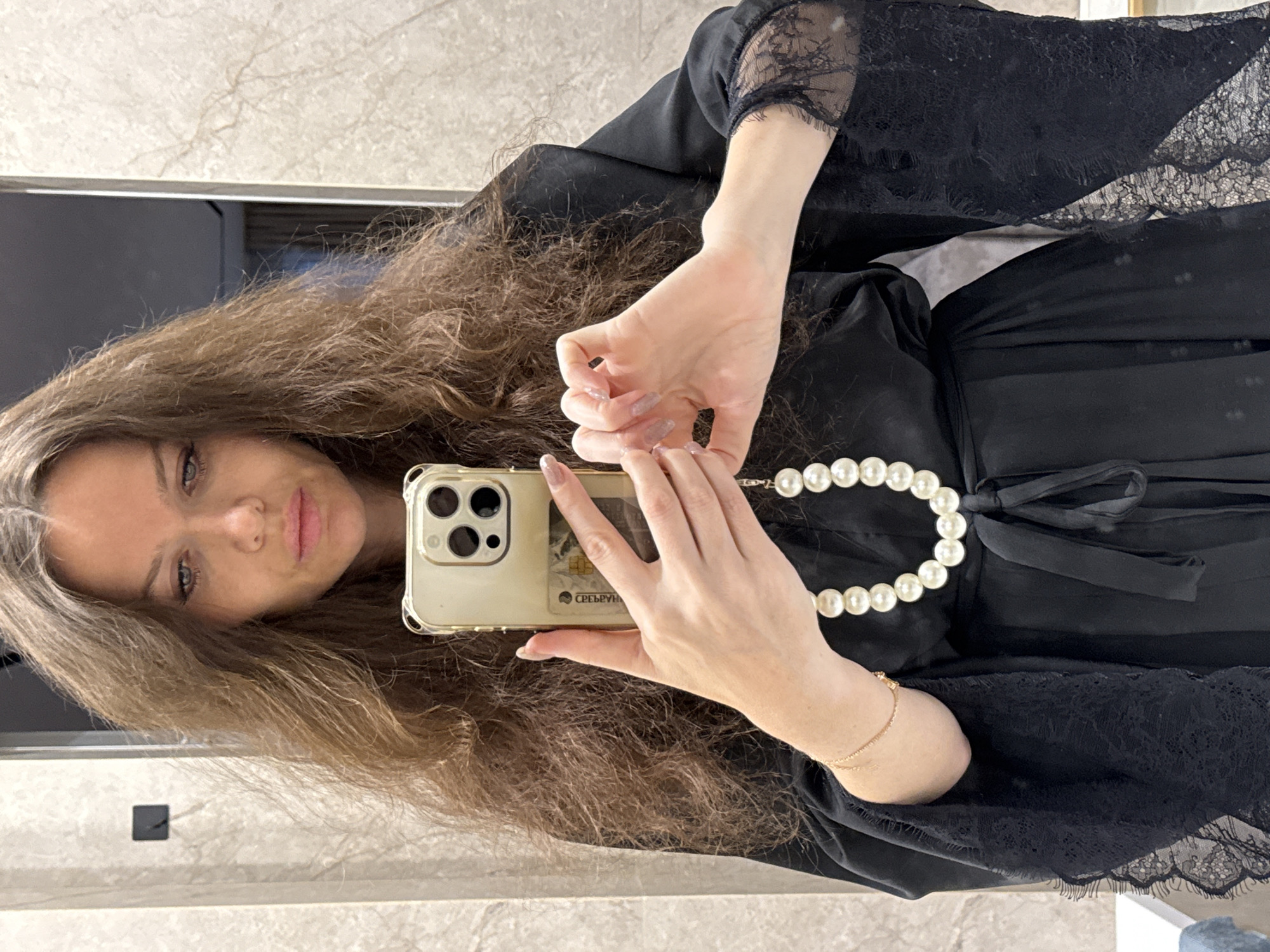 A woman with long brown hair is taking a selfie in a bathroom mirror. She is holding an iPhone in front of her, showing the phone case and a credit card. She is wearing a black robe with lace trim, tied with a satin ribbon, and a pearl necklace. A thin gold bracelet is on her wrist. The bathroom has light-colored tile walls and a dark rectangle on the wall near the bottom left corner. The credit card shows "СБЕРБАНК" which translates to Sberbank in English, a Russian bank, which would indicate the location is most likely in Russia. Without further evidence, the city cannot be determined, but Moscow, Russia is a possible location given the prevalence of Sberbank in the country.