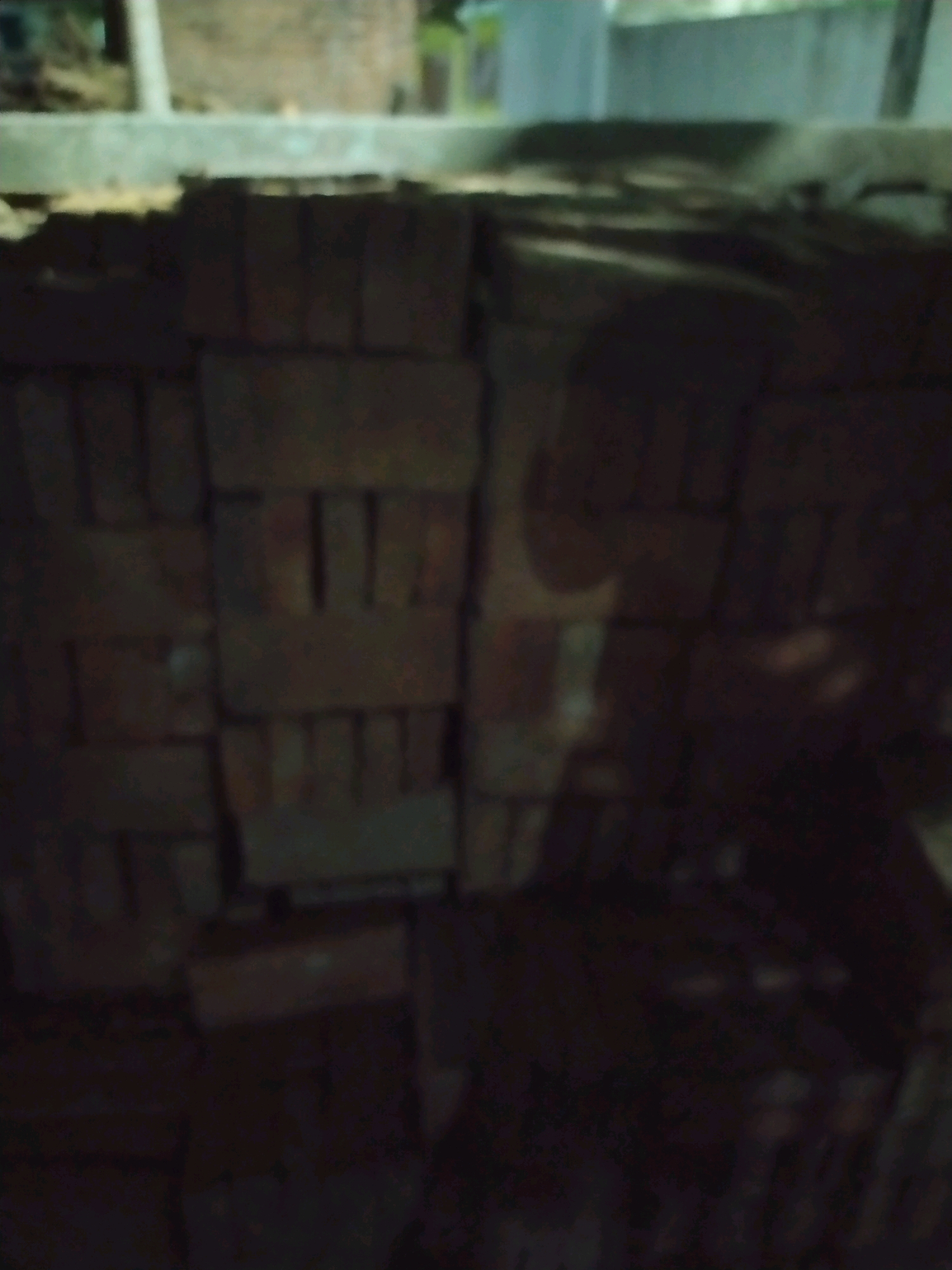 The image depicts a stack of bricks in a dimly lit outdoor environment. The bricks appear to be made of clay and are stacked in a rough, orderly fashion. The background is out of focus but suggests a building or wall behind the bricks, with some vegetation visible. The lighting is poor, casting deep shadows and making it difficult to discern fine details. There are no people or animals visible in the frame. The scene appears to be located in an industrial or construction area, possibly a storage yard for building materials. The overall impression is one of a functional, utilitarian space.