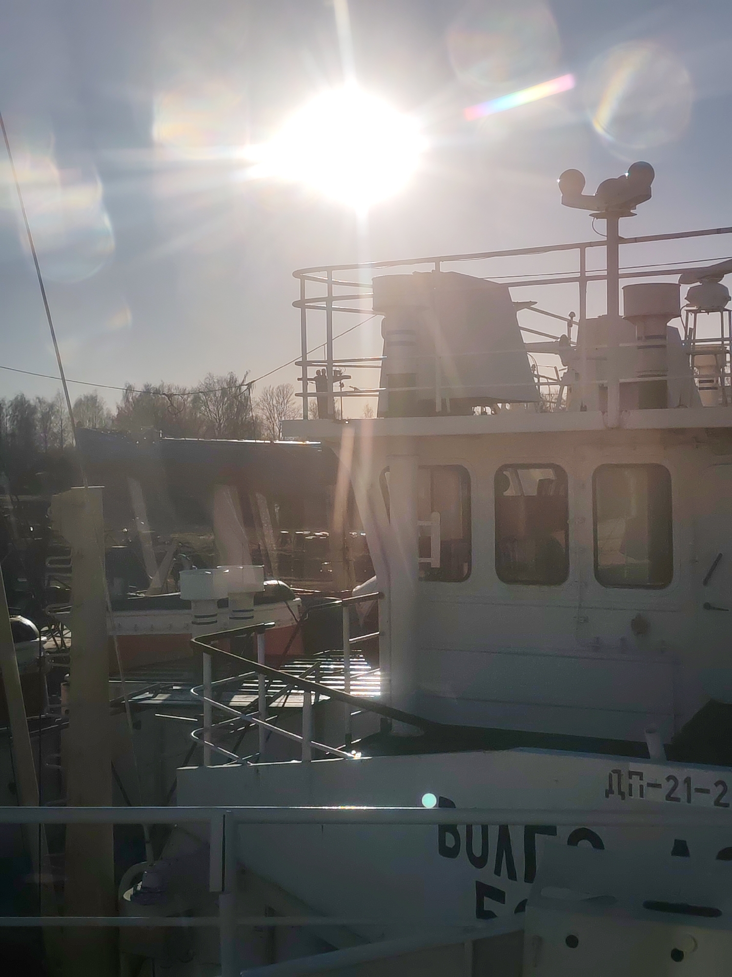 The image captures a sun-drenched view of a white vessel, likely a workboat or small ship, docked in what appears to be a shipyard or port. The sun is bright and low in the sky, positioned in the upper right quadrant, creating intense lens flare and light streaks across the frame, backlighting the scene.
The main subject is the superstructure of the white boat, featuring multiple windows, railings, and what seem to be radar or communication equipment masts on its top deck. The boat has a utilitarian appearance, with visible details of its construction and navigation elements. In the mid-ground to the left, parts of another, darker vessel are visible, suggesting a busy docking area. Further in the background, a line of bare trees hints at a temperate climate, possibly late autumn or early spring. The sky is clear and bright blue.
No specific human activity or event is clearly depicted; the scene gives the impression of a vessel at rest or undergoing maintenance. A faint silhouette of a person might be discernible inside one of the boat's windows.
Notable text is visible on the side of the main boat: "ДП-21-2" (DP-21-2) is clearly legible. Below that, the word "ВОЛГА" (VOLGA) is partially visible, with "ВОЛГ" and part of the "А" being discernible before the text is cut off or obscured. The bright sun and lens flares are prominent features, contributing to the ethereal, almost dreamlike quality of the light.