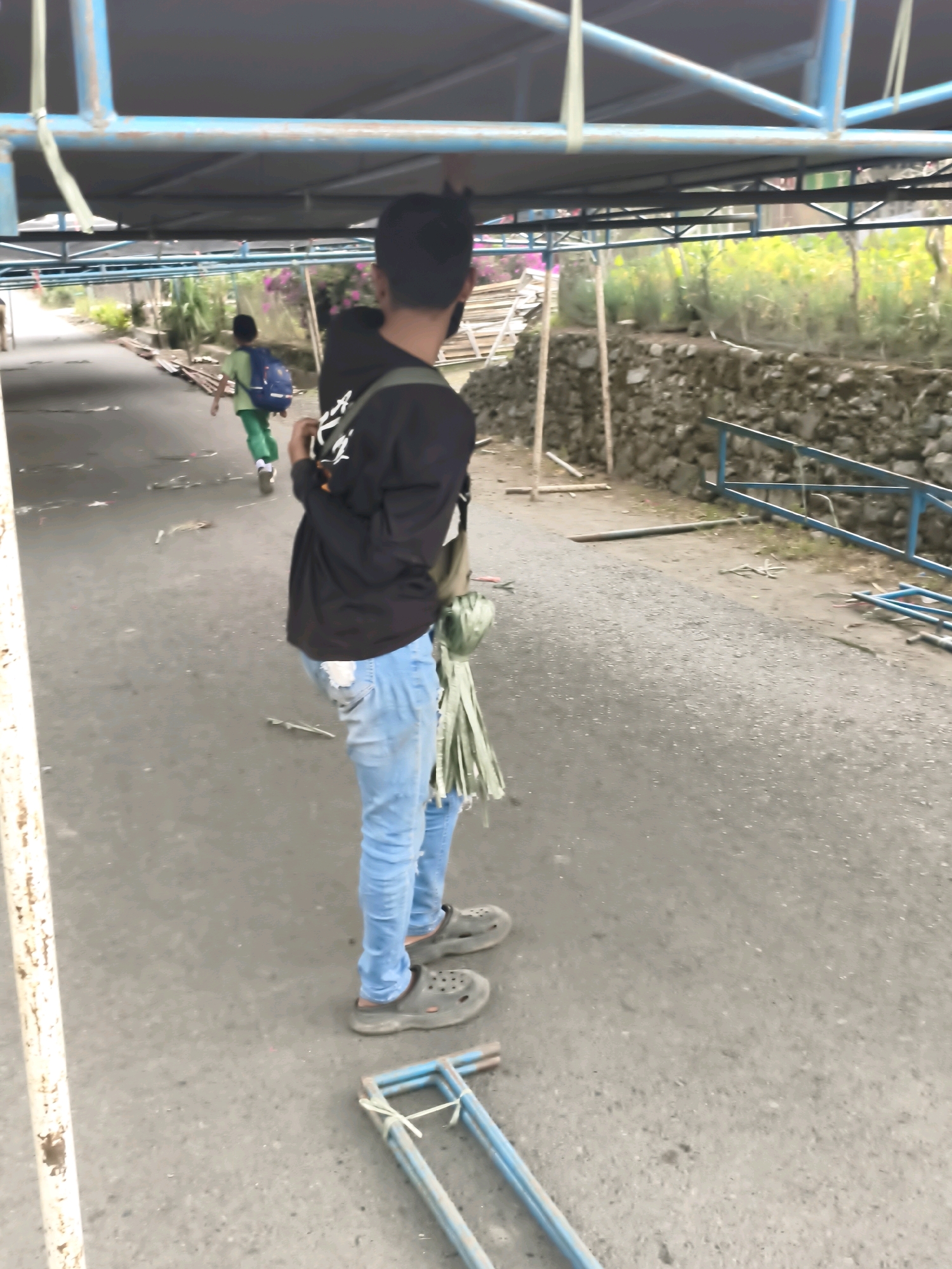 In Lindung Jaya, Indonesia, a person stands on an asphalt surface beneath a blue metal structure with a dark gray tarp. The individual, facing away from the camera, wears ripped light blue jeans and dark gray slip-on shoes. They have a black long-sleeved shirt and a green backpack. In the background, another person wearing a green shirt and pants with a blue backpack is walking away from the camera. The environment includes a stone wall, greenery, and scattered debris on the ground.