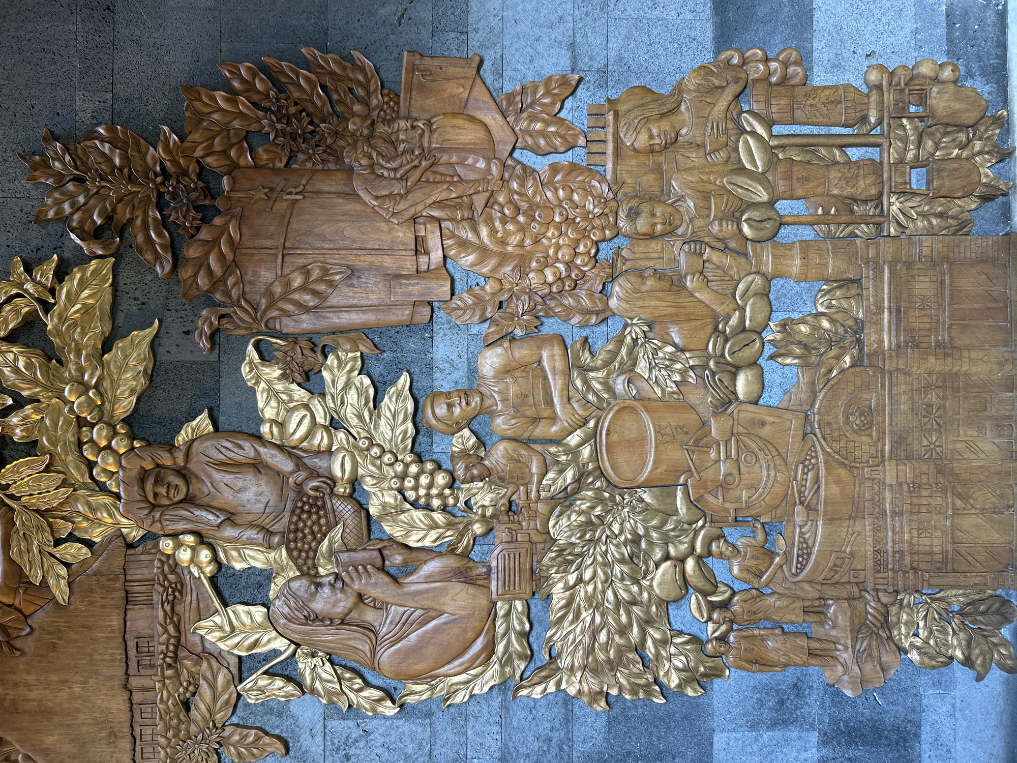 This is a detailed, close-up, eye-level shot of a large, intricate wooden relief carving mounted on a dark, textured wall, likely in Kuta, Indonesia, given the cultural context suggested by the artwork. The carving depicts scenes related to coffee cultivation and consumption, rendered in warm, natural wood tones, with some elements highlighted in gold.
The artwork is densely packed with figures and motifs. In the upper section, a figure is shown carrying a basket of coffee beans on their head, with a rustic structure in the background. Below this, a woman with long hair drinks from a mug, surrounded by coffee leaves and berries. In the middle section, several figures appear to be working with coffee beans and machinery, including one person operating a grinding mechanism. To the right, a large wooden barrel or vat is depicted, adorned with coffee flowers and leaves, and a Starbucks logo is subtly visible.
The lower part of the carving features a group of people, some holding cups, suggesting they are enjoying coffee. Various coffee-related objects are also present, such as large coffee beans, processing equipment, and sacks. The overall composition tells a narrative of the coffee journey from bean to cup, employing a realistic yet stylized approach to the human figures and natural elements. The dark, stone-like wall provides a stark contrast to the warm wood and gold of the carving, making the artwork the clear focal point.