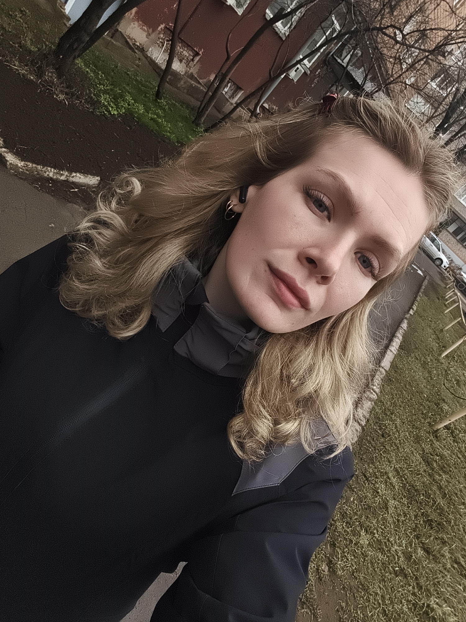 The image depicts a woman outdoors in what appears to be Izhevsk, Russia, based on architectural cues. She is wearing a dark jacket and has blonde, wavy hair. She has an earbud in her left ear and is wearing earrings. The background includes trees without leaves, a building with red brick on the facade, and patches of grass. There is a walkway or pavement to the left of the woman. A vehicle is partially visible in the background.