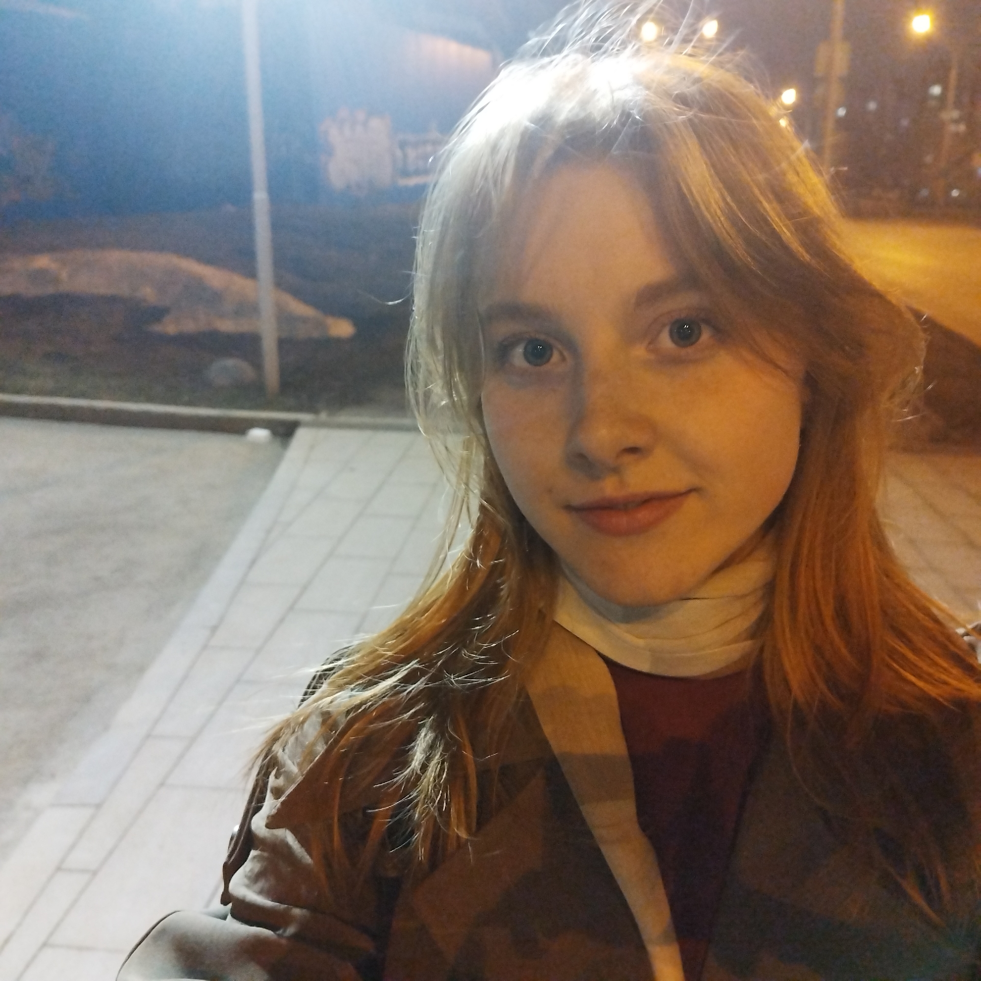 A selfie taken at night shows a fair-skinned young woman with strawberry blonde hair, blue eyes, and freckles. She wears a dark-colored shirt under a brown jacket or coat, and a white scarf is around her neck. The background indicates an urban environment at night. The setting is illuminated by streetlights and artificial light. Paved ground and a curb are visible beneath her. In the background, there's a building or structure possibly with some graffiti. Given the prompt context, the location is potentially in Yekaterinburg, Russia, although specific landmarks aren't identifiable in the image.