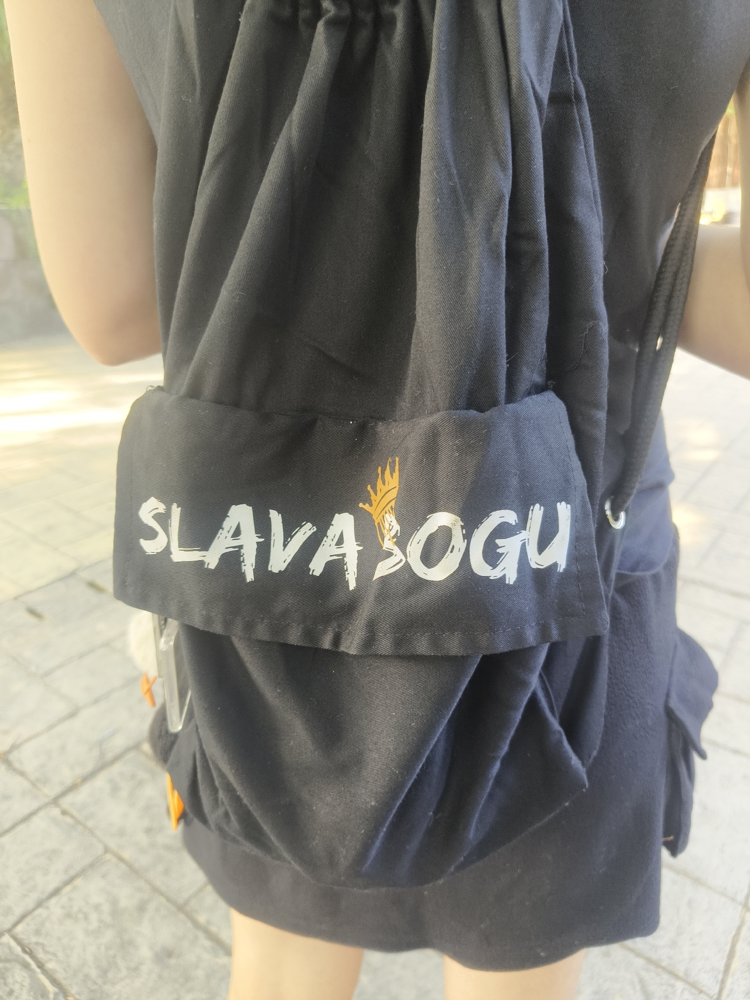 A person is viewed from the back, wearing a black backpack with the word "SLAVA SOGU" and a golden crown logo printed on a white band across the front. The person is standing on a paved area with sunlight reflecting off the ground, suggesting an outdoor location. The background is blurred, but appears to be a park or recreational area with some greenery visible. The setting is consistent with an urban park in Kuta Selatan, Indonesia.