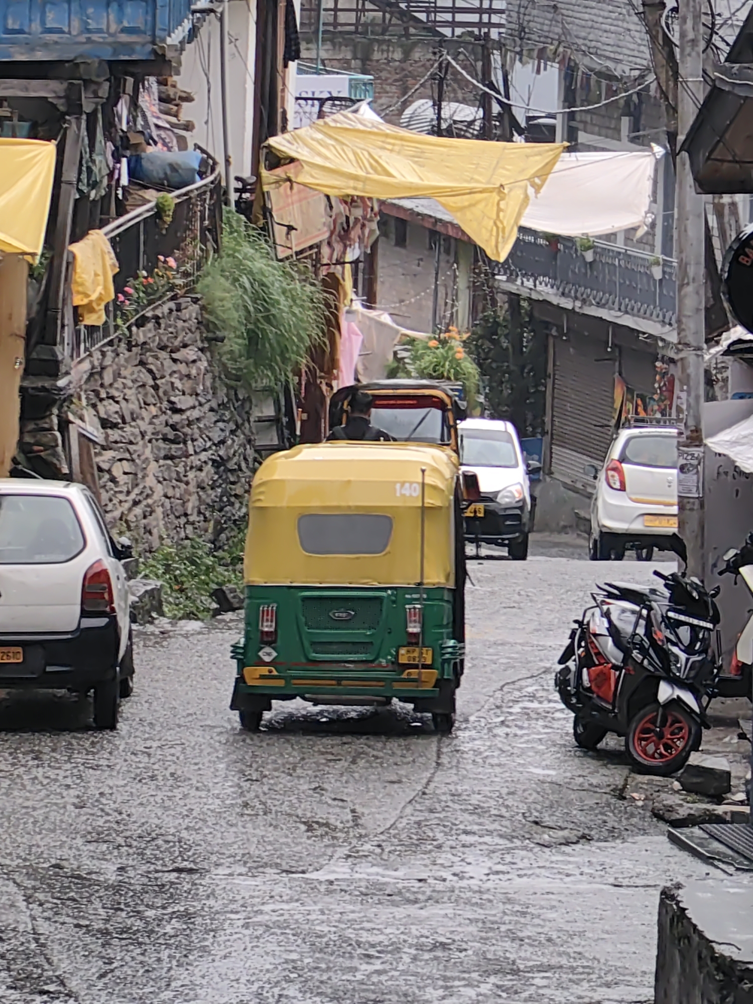 The image depicts a narrow, wet street in Vashisht, India. An auto-rickshaw, predominantly yellow and green, is the central focus, moving away from the camera. To its left, a white car is parked. To the right of the auto-rickshaw, a white car is also parked, and further back on the right, a motorcycle with red wheels is visible. The street is paved with an uneven surface, wet and reflective, indicating recent rain or moisture. Buildings line both sides of the street, with some featuring balconies and awnings, including yellow tarpaulins. Clothing and other items are visible hanging on railings and structures along the buildings. The scene appears to be a typical street in a populated urban or semi-urban area, with signs of daily life and activity.