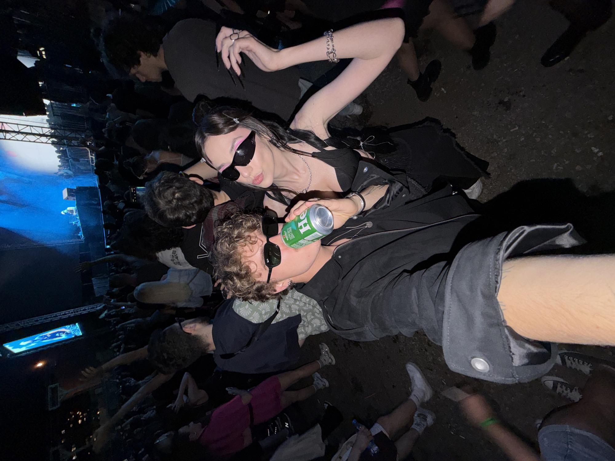 The image captures a low-angle selfie taken at an outdoor music festival. In the foreground, a young man with curly blonde hair and sunglasses is drinking from a can of Heineken. Beside him, a young woman with dark hair styled in pigtails, also wearing sunglasses, poses with her hand raised and holding the can. Both are dressed in dark clothing.
Behind them, a crowd of people is visible, many with their hands raised, suggesting they are enjoying the performance. A stage with a DJ and large screens displaying blue light is visible in the background, indicating the presence of a live event. The ground is covered in gravel. The overall atmosphere appears to be energetic and celebratory, consistent with a music festival environment. The location is not specified to be in Palilula Urban Municipality, Serbia.