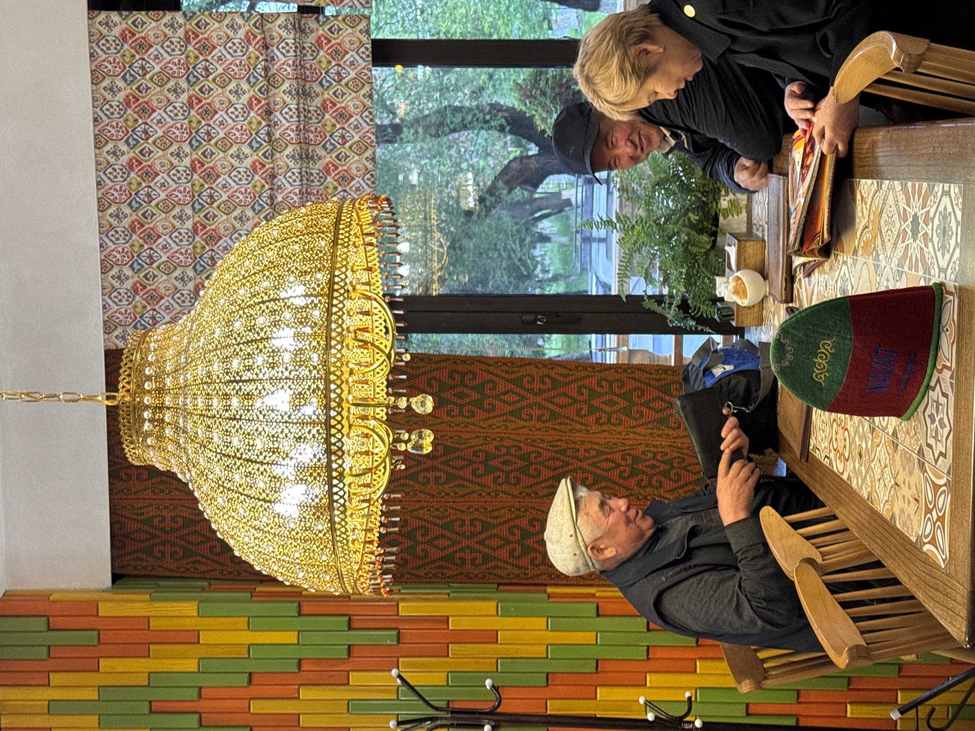 This media file captures an indoor scene, likely a restaurant or cafe in Bishkek City, Kyrgyzstan. Three individuals are seated around a wooden table with a distinctive tiled surface.
In the foreground, an older man wearing a light-colored flat cap and a dark jacket is engrossed in a device held in his hands. Across the table, a woman with short blonde hair and a man in a dark cap are reviewing menus; the man in the cap is smiling, suggesting a pleasant atmosphere.
The setting is richly decorated. A prominent, ornate golden chandelier with intricate filigree work and numerous lights hangs from the ceiling. The walls feature a colorful, stacked brick-like pattern in green, yellow, and orange, topped by a section of patterned fabric or wallpaper with geometric designs in reddish-brown, green, and orange. A large window in the background provides a view of lush green trees, indicating an outdoor presence and natural light, likely daytime.
On the table, alongside the menus, is a distinctive green and red traditional-looking hat or cap, possibly a Kyrgyz Ak-Kalpak, with "AK KANAT" visible in white text on its red portion. A potted fern and a white decorative object also adorn the table. The scene evokes a comfortable and culturally infused dining experience.