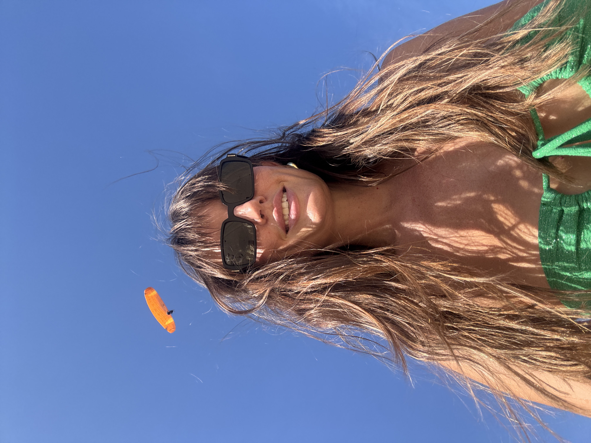 A woman wearing sunglasses and a green top looks up at a clear blue sky where a paraglider is visible. The location is likely the city of Kuta Selatan, Indonesia.