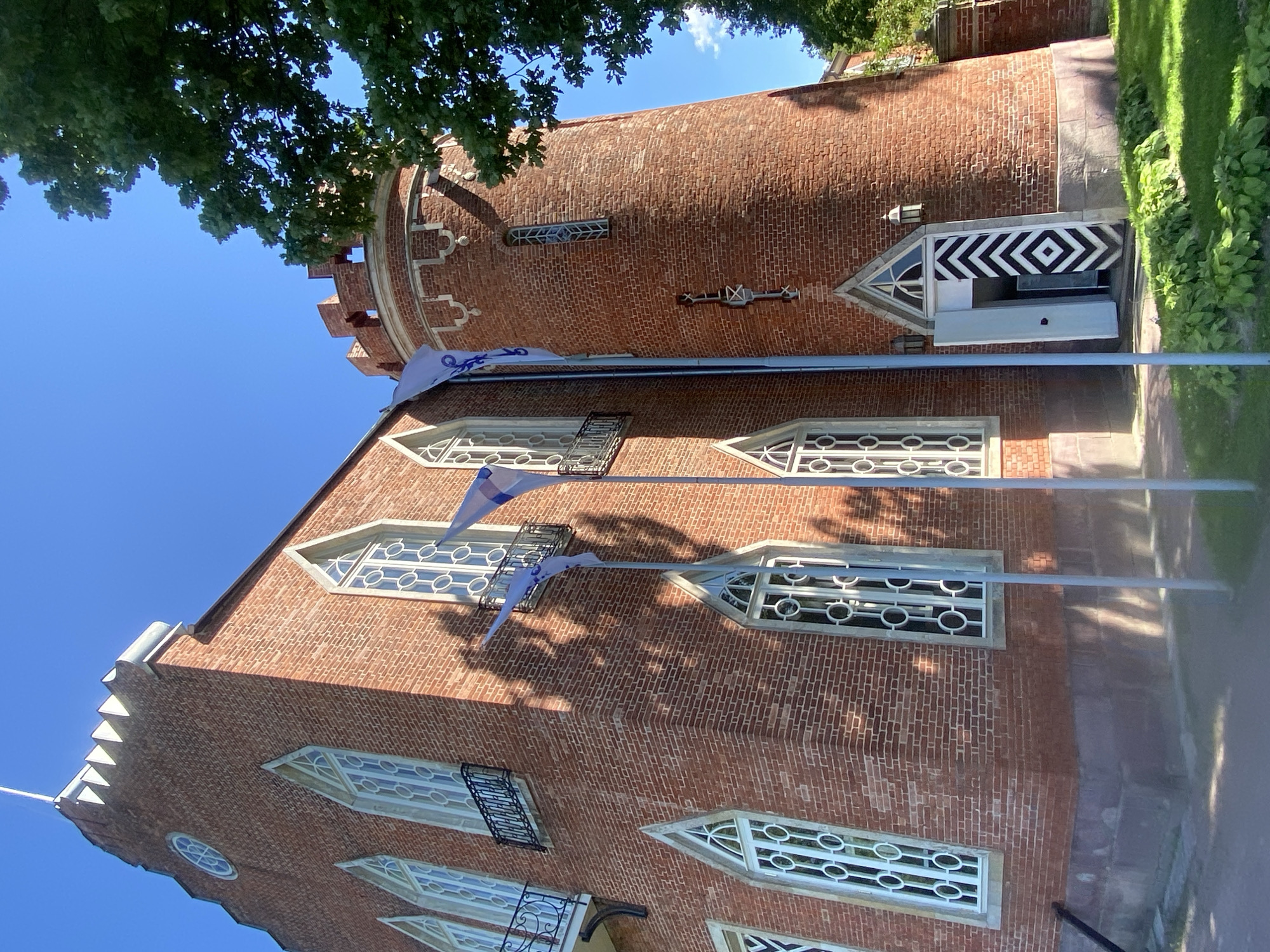 The image shows a brick building with a rounded tower, featuring Gothic-style windows with circular patterns. Several flags are planted in front of the building. The building is situated in a grassy area with trees. The scene is illuminated by bright sunlight, casting shadows on the building's facade. A close-up of the entrance reveals a doorway with black and white diagonal stripes on the doorjamb. This building is identified as the Arsenal in the city of Pushkin, Russia.
