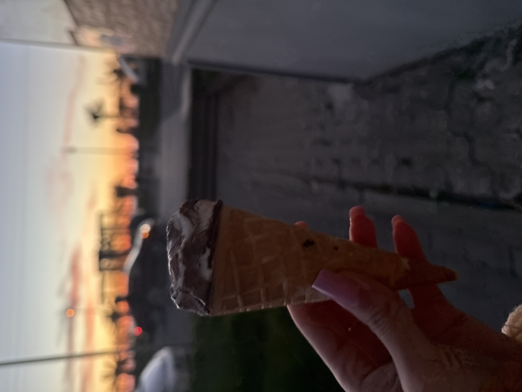 The photo shows a hand holding an ice cream cone. The ice cream is two-toned, possibly chocolate and vanilla. The background shows a sunset scene with what appear to be palm trees, suggesting a tropical or coastal location. The image appears to have been taken outside, possibly on a balcony or near a building's exterior wall. Based on the palm trees and sunset, the location is likely Batumi, Georgia.