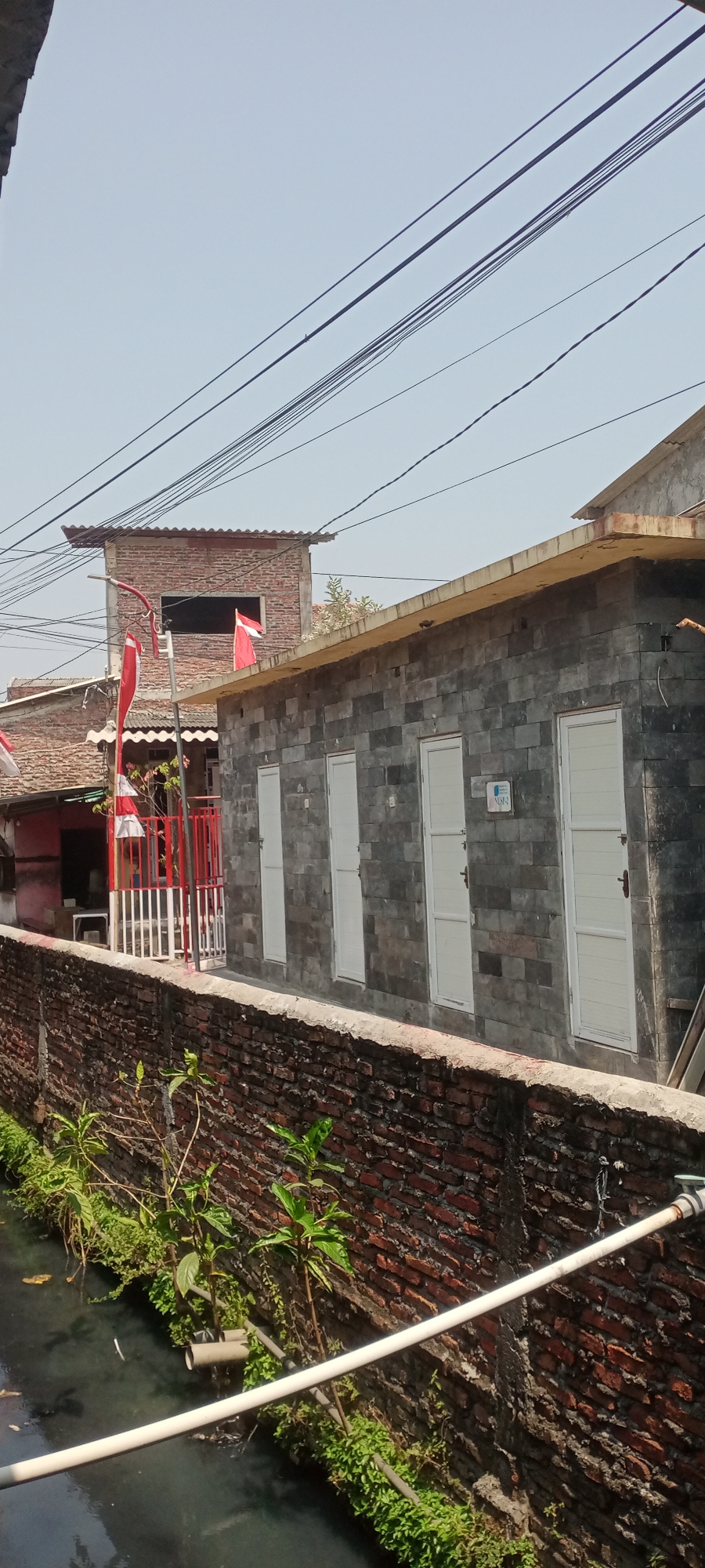 The image depicts an outdoor scene in the city of Semarang, Indonesia, likely in a residential area with a narrow canal running along a brick wall. The canal has murky water and is lined with green vegetation and some discarded pipes. A prominent brick wall, weathered and aged, runs along the canal.
Adjacent to the wall is a building constructed with dark gray stone blocks, featuring several identical white doors. Above this building, a portion of another structure made of red bricks is visible, topped with a corrugated metal roof. This brick structure has a large, dark rectangular opening resembling a window. Two Indonesian flags, red and white, are displayed on poles near the brick structure.
Further to the left, another red-bricked building with a tiled roof is visible, partially obscured by the central stone building and flags. A red and white striped railing encloses a small elevated area in front of this red-bricked building. Electrical wires are strung across the sky, indicating the presence of infrastructure. The sky is clear and pale blue, suggesting it is daytime. No people are visible in the image. The overall impression is a view of urban housing and infrastructure in Semarang.
