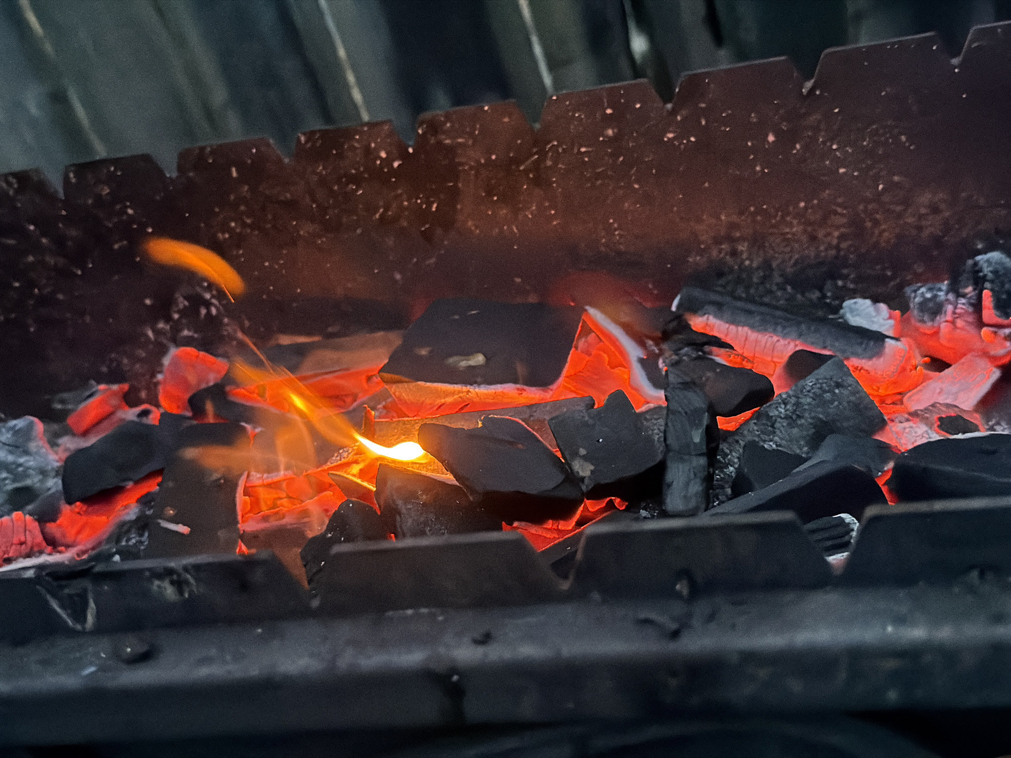 The image shows burning charcoal inside a metal brazier. The charcoal is glowing red-orange in the center, with dark black pieces surrounding it. Flames are visible rising from the hot coals. The brazier has a notched edge. The overall setting appears to be low-light. There is no information to support that this image was taken in Ol'khovo, Russia.