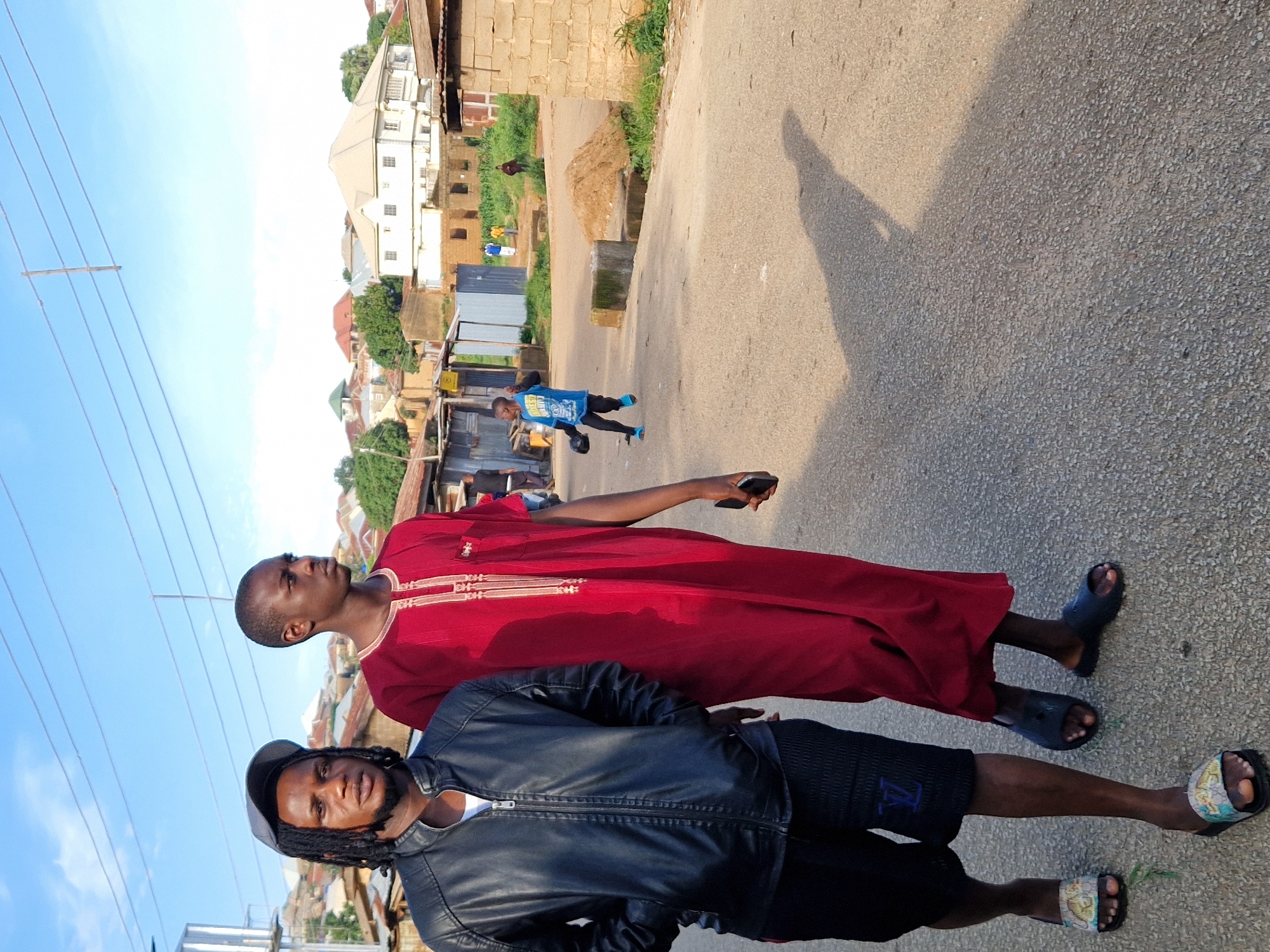 Two young men are walking down a paved street in Kabong, Nigeria. The man on the left wears a black leather jacket, black shorts with a blue logo, and a grey baseball cap. He has dreadlocks and is looking forward. The man on the right wears a dark red robe with gold trim and is also looking forward. He holds a dark-colored smartphone in his right hand. In the background, there are several buildings with corrugated metal roofs and some taller, more modern houses. A young boy in a blue t-shirt and black pants runs in the middle ground, casting a shadow. Power lines are visible in the sky, along with scattered clouds. The scene appears to be during daylight, with long shadows cast on the street.