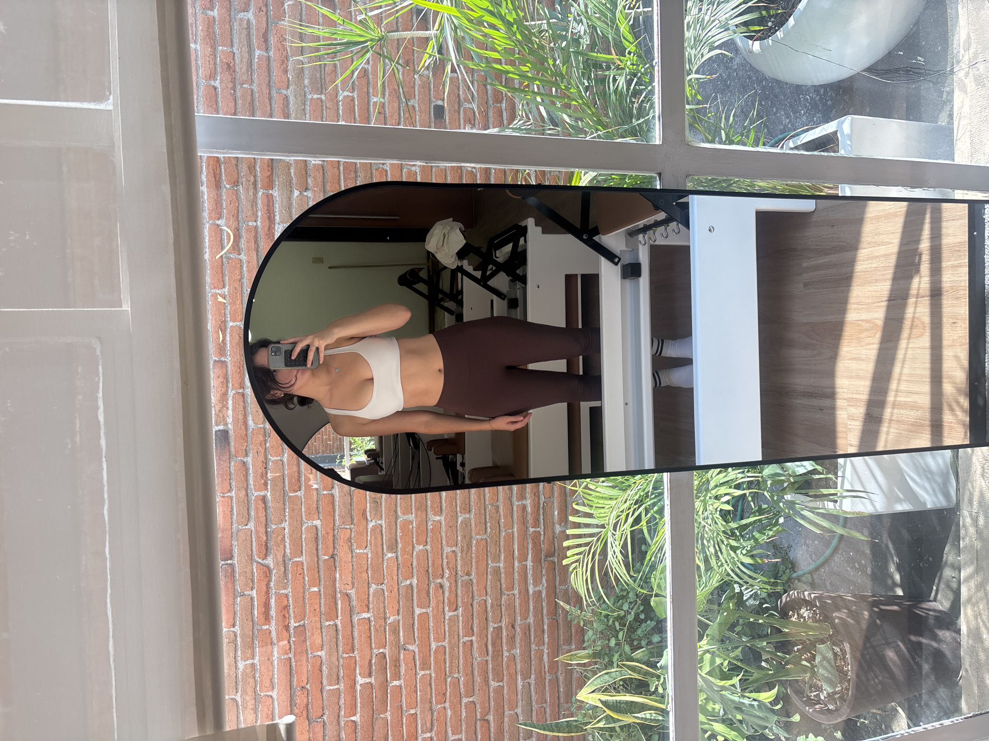 The image shows the reflection of a woman in a mirror. She is wearing a white top and brown leggings, and she is holding a smartphone in her left hand to take the picture. The mirror is positioned in front of a brick wall and a window through which greenery is visible. Inside the room there is pilates equipment, with a white towel hanging from the frame. Based on the architectural style, the presence of red brick, and the lush green vegetation, the image was likely taken in the city of Puebla, Mexico.
