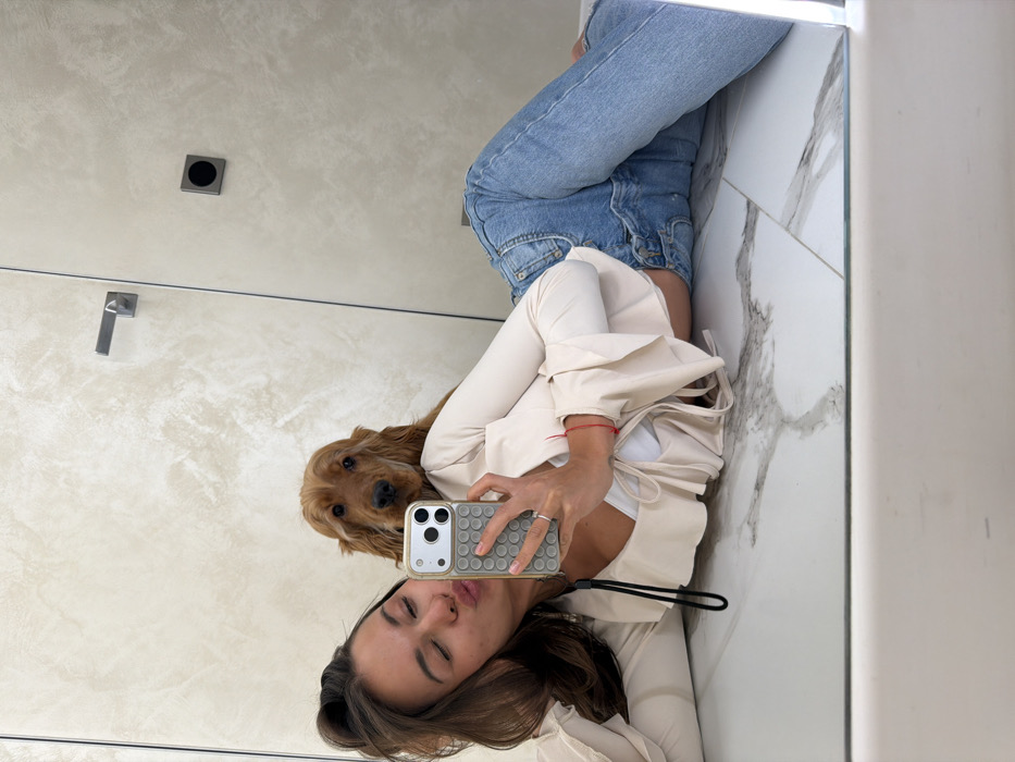The image displays a mirror reflection of an indoor scene. A young adult female with dark hair is lying on her left side on a light-colored, marble-patterned tiled floor. She wears light blue denim jeans and a long-sleeved, ruffled, light-beige crop top. Her left arm is bent, holding a smartphone with a distinct grey "pop-it" style case and a gold-colored frame. She is taking a selfie, with her eyes closed and lips pursed as if blowing a kiss. A thin red string bracelet is visible on her left wrist, and a ring is on her right-hand ring finger. A black lanyard hangs from the phone case. Immediately behind and above her, an English Cocker Spaniel-type dog with golden-brown fur rests its head on her shoulder/upper back, looking directly into the mirror. The background, visible in the mirror, consists of light-beige, textured walls. A modern, metallic horizontal door handle is visible on the left, and a square-shaped dark control panel or switch is mounted on the wall further right. The specific location within the city of Krasnoyarsk, Russia, cannot be determined from the visual information provided.