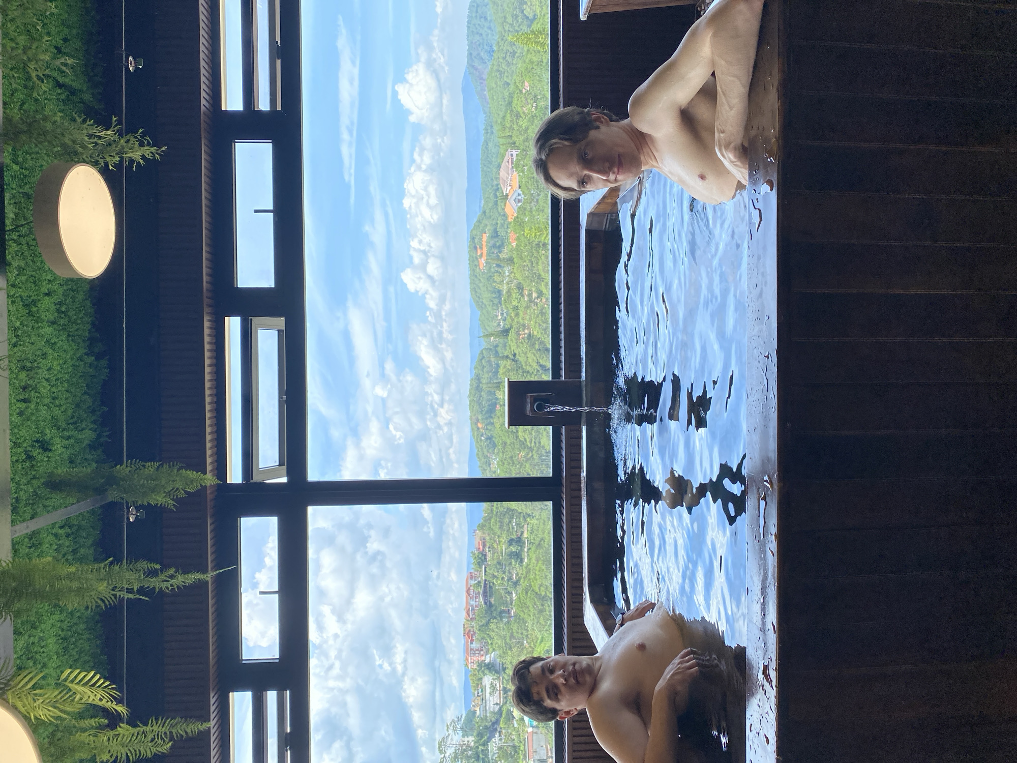Two shirtless young men are relaxing in a large wooden hot tub. The hot tub is situated indoors, near a large window with a view of a city nestled in a lush, green valley. The window overlooks a cityscape with red-roofed buildings and rolling hills covered in trees, characteristic of Da Lat, Vietnam. Water is flowing into the tub from a dark-colored fountain fixture. Hanging plants and artificial greenery are visible on the ceiling above the tub, and a modern light fixture hangs to the left. The water in the tub is clear and reflects the light. Both men are looking towards the camera.