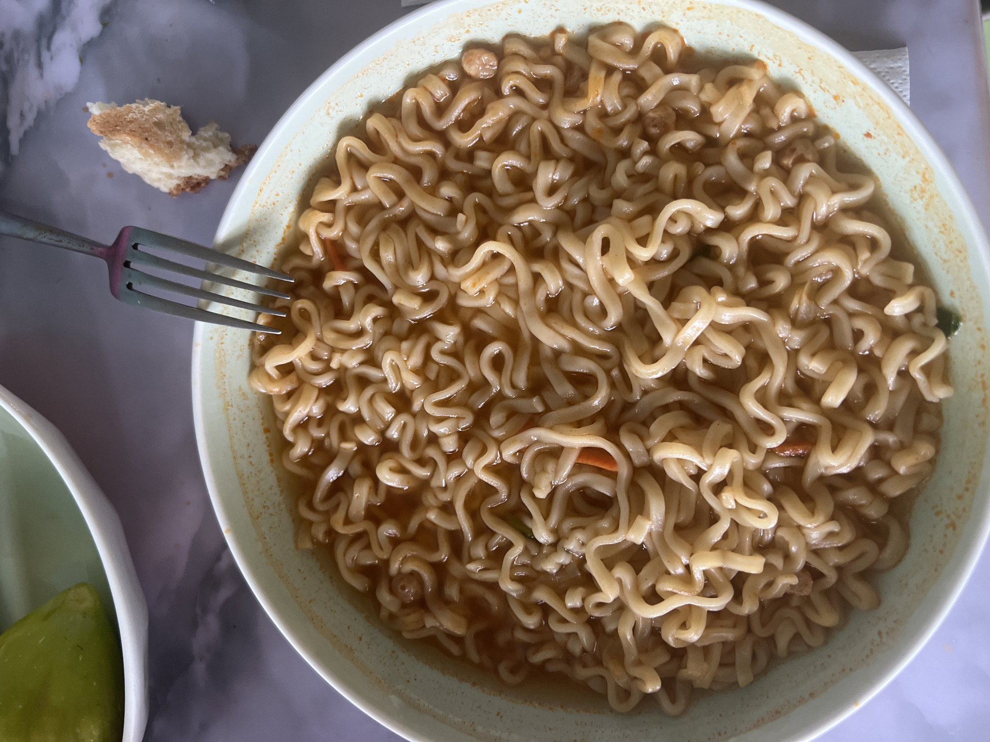 A close-up, overhead shot shows a bowl of instant ramen noodles with broth. The noodles are wavy and golden brown, mixed with small pieces of orange and green vegetables. A silver fork rests on the edge of the bowl, its tines submerged in the broth. To the right of the bowl, a small, torn piece of white bread with a browned crust lies on a patterned surface. In the background, out of focus, a portion of another bowl containing a green object, possibly a vegetable, is visible. The scene is brightly lit, suggesting an indoor setting, possibly a kitchen or dining area. The surface beneath the bowl appears to be a marble-patterned countertop. This image does not contain information about the city of Şamaldı-Say, Kyrgyzstan.