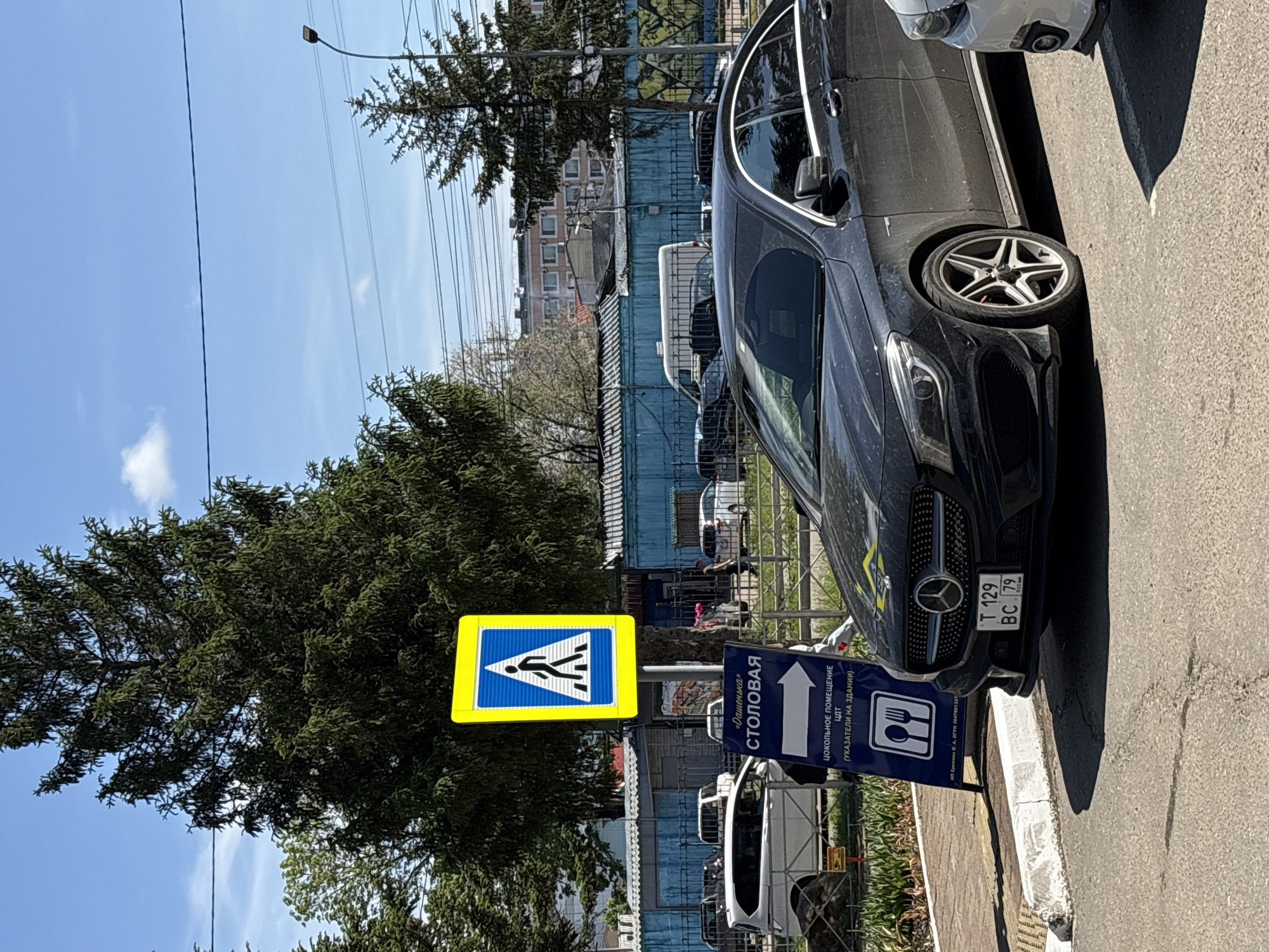 A black Mercedes-Benz is parked on an asphalt road in front of a blue fence and a sidewalk. A blue sign with white text and symbols is placed on a stand to the left of the car. The sign has a yellow pedestrian crossing symbol at the top and below it, text that reads "«Дашенька» СТОЛОВАЯ" and an arrow pointing to the right. Further down, there is a symbol of cutlery and text reading "ЦОКОЛЬНОЕ ПОМЕЩЕНИЕ ЦДТ (УКАЗАТЕЛИ НА ЗДАНИИ)". The license plate of the Mercedes-Benz reads "T 129 BC 79". In the background, there are trees, buildings, and power lines under a clear blue sky. The scene appears to be a street in Birobidzhan, Russia, likely near a dining establishment or community center.