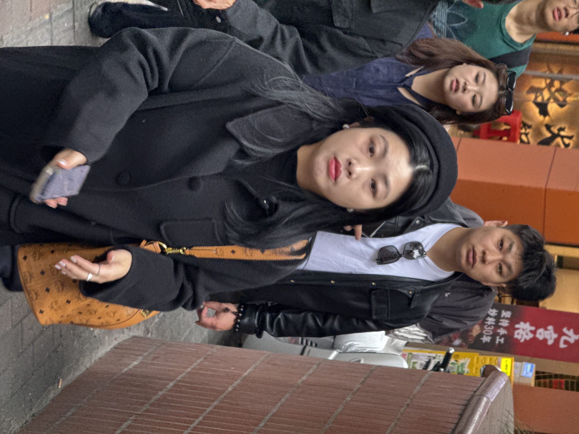 The image captures a crowded urban street scene in Yuzhong District, China, depicting multiple pedestrians moving along a paved walkway.
In the immediate foreground, a young East Asian woman, approximately 20-30 years old, is centered. She wears a black beret, a black long-sleeved overcoat with large buttons, and a black crew-neck top. Her dark, straight hair falls over her shoulders. She has noticeable red lipstick, subtle eye makeup, and small white stud earrings. Her right hand, featuring light-colored nail polish, holds a grey smartphone. A tan-brown shoulder bag with a repeating dark monogram pattern and gold-colored hardware hangs from her left shoulder. Her expression is neutral, and she appears to be walking forward.
Behind her and to the right, a young East Asian man, also 20-30 years old, walks. He wears a white t-shirt, a black leather-like jacket, and dark sunglasses are hooked onto his t-shirt collar. A dark beaded bracelet is visible on his left wrist. His gaze is directed forward.
To the left of the man and slightly behind the foreground woman, another young East Asian woman, 20-30 years old, is visible. She has shoulder-length brown hair with dark sunglasses resting on top, and wears a dark blue top. Her mouth is slightly open, and she looks ahead. Additional figures are partially visible further in the background, including a person in a green t-shirt to the far left.
The environment features a reddish-brown brick wall on the right side of the frame. To the left, a building with an orange-brown facade displays a large, stylized Chinese character, possibly "火" (fire), in a metallic finish. Further back on the left, a vertical red sign with white Chinese characters reads "九宫格" (Jiugongge, a type of hotpot) and includes smaller text stating "坚持手工炒料30年" (persistently handmade stir-fried ingredients for 30 years). The ground is paved with grey interlocking stones. The scene suggests a busy commercial or pedestrian area during daylight hours.