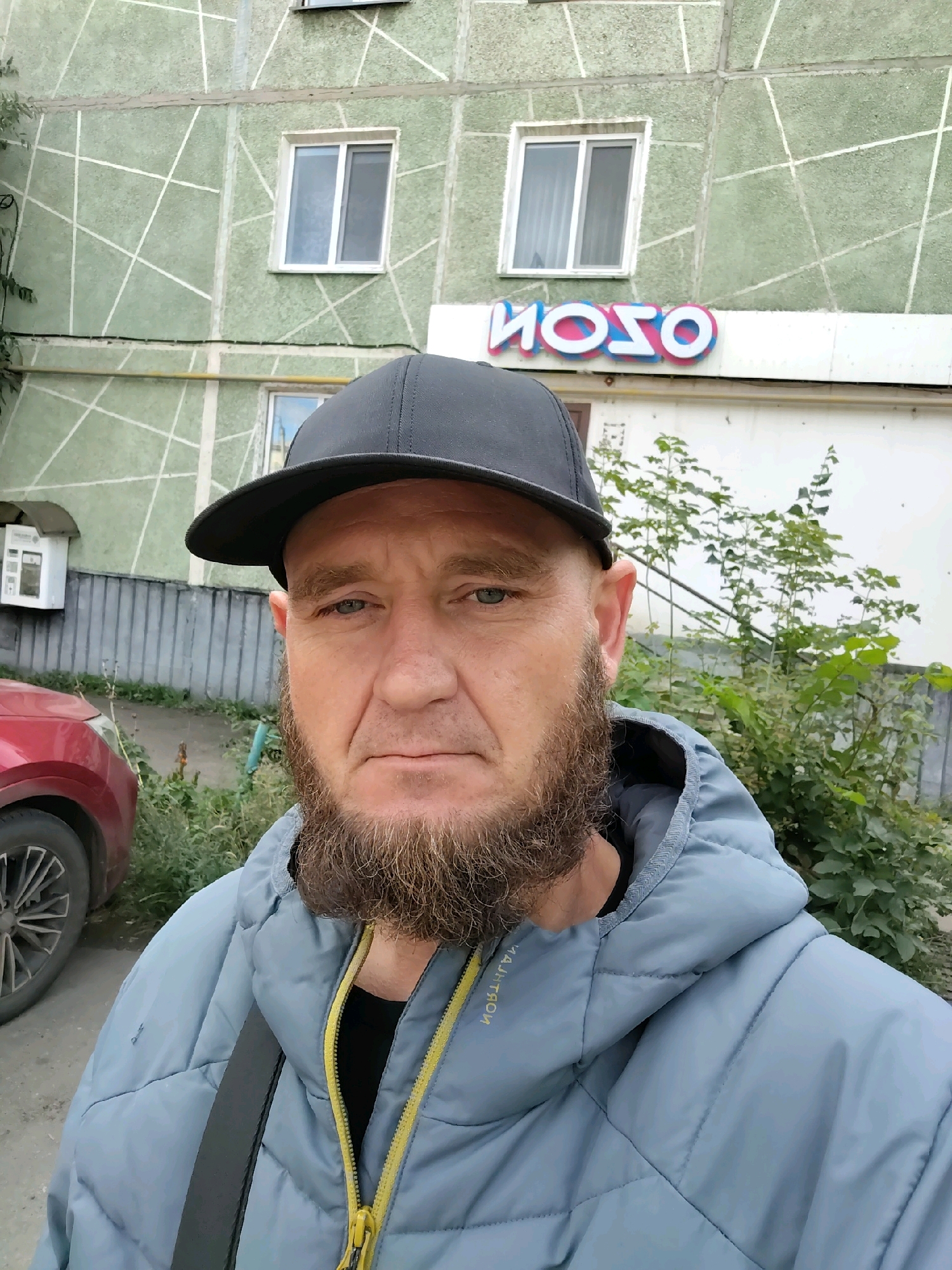 A man with a beard wearing a black baseball cap and a light blue hooded jacket is standing outside a building in the city of Petropavl, Kazakhstan. The building has a light green facade with a geometric white pattern. There are two windows on the upper part of the building. Below the windows, a sign with colorful letters reads "HOSO". A red car is partially visible on the left side of the image, parked next to the building. Green foliage and bushes are present around the building and the car. The man is looking towards the camera.