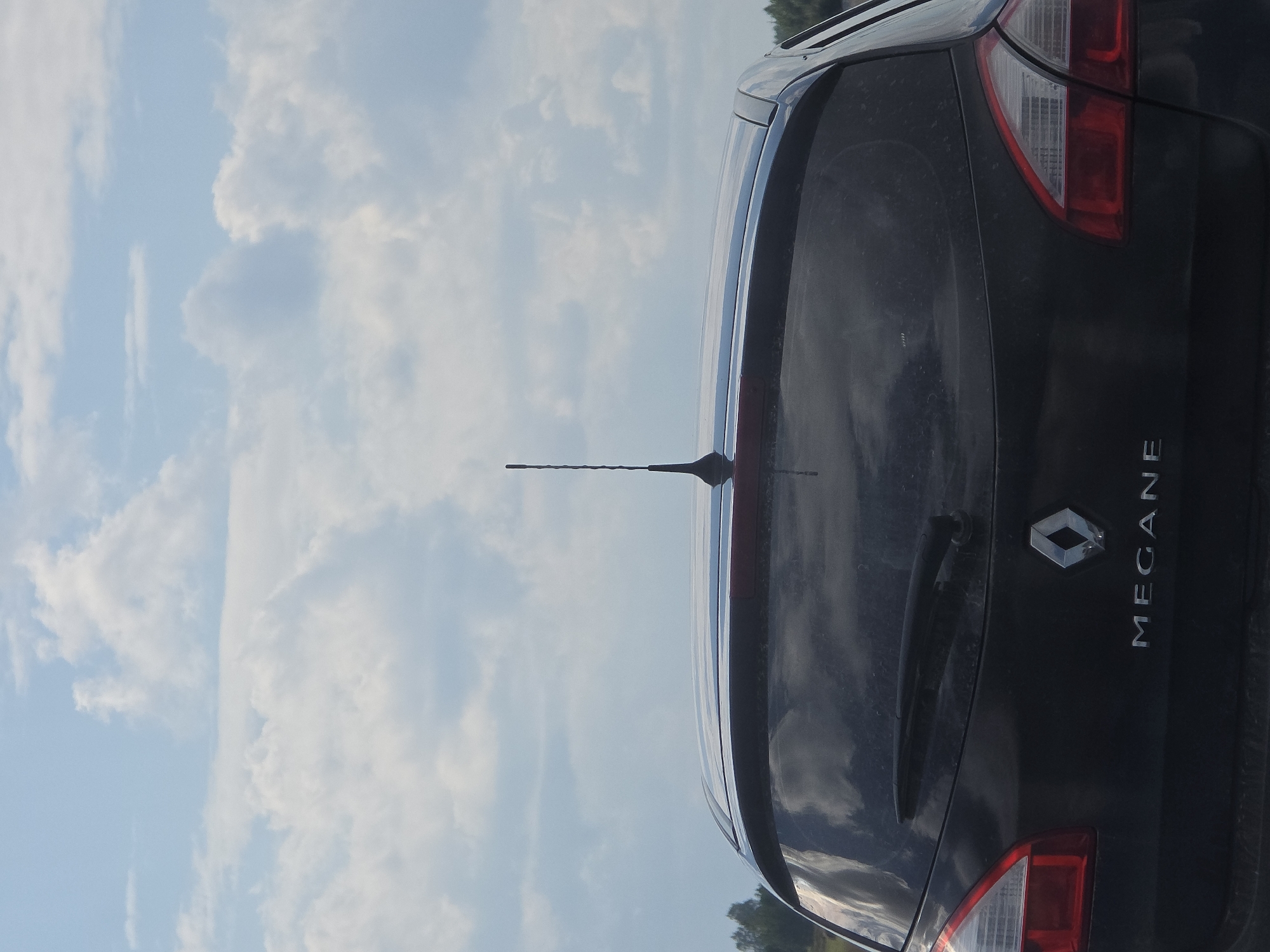 The image shows the rear of a black Renault Megane hatchback parked outdoors. The car has a rear windshield wiper and an antenna on the roof. The car's emblem and "MEGANE" badging are visible on the trunk. The background consists of a blue sky with scattered white clouds and a faint treeline in the distance. The image quality is moderate, with some dust or dirt visible on the car's surface. The location is identified as Yaltyrova, Russia. There are no people or animals visible in the image, and no action is taking place. The scene is a static, outdoor shot of a parked vehicle.