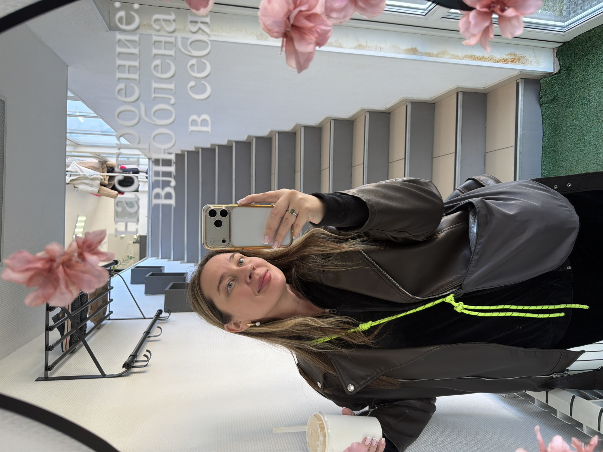 A woman is taking a selfie in a mirror. She is wearing a brown leather jacket over a black top with a neon yellow drawstring. She holds a white disposable cup with a straw in her left hand. She is holding a phone with a gold-colored case in her right hand.
The mirror reflects a staircase, a coat rack with shoes and coats, and Russian text on the wall. Pink flowers are attached to the mirror. The setting is likely inside a building in the city of Chaikovsky, Russia. A patch of artificial grass is also visible in the reflection.