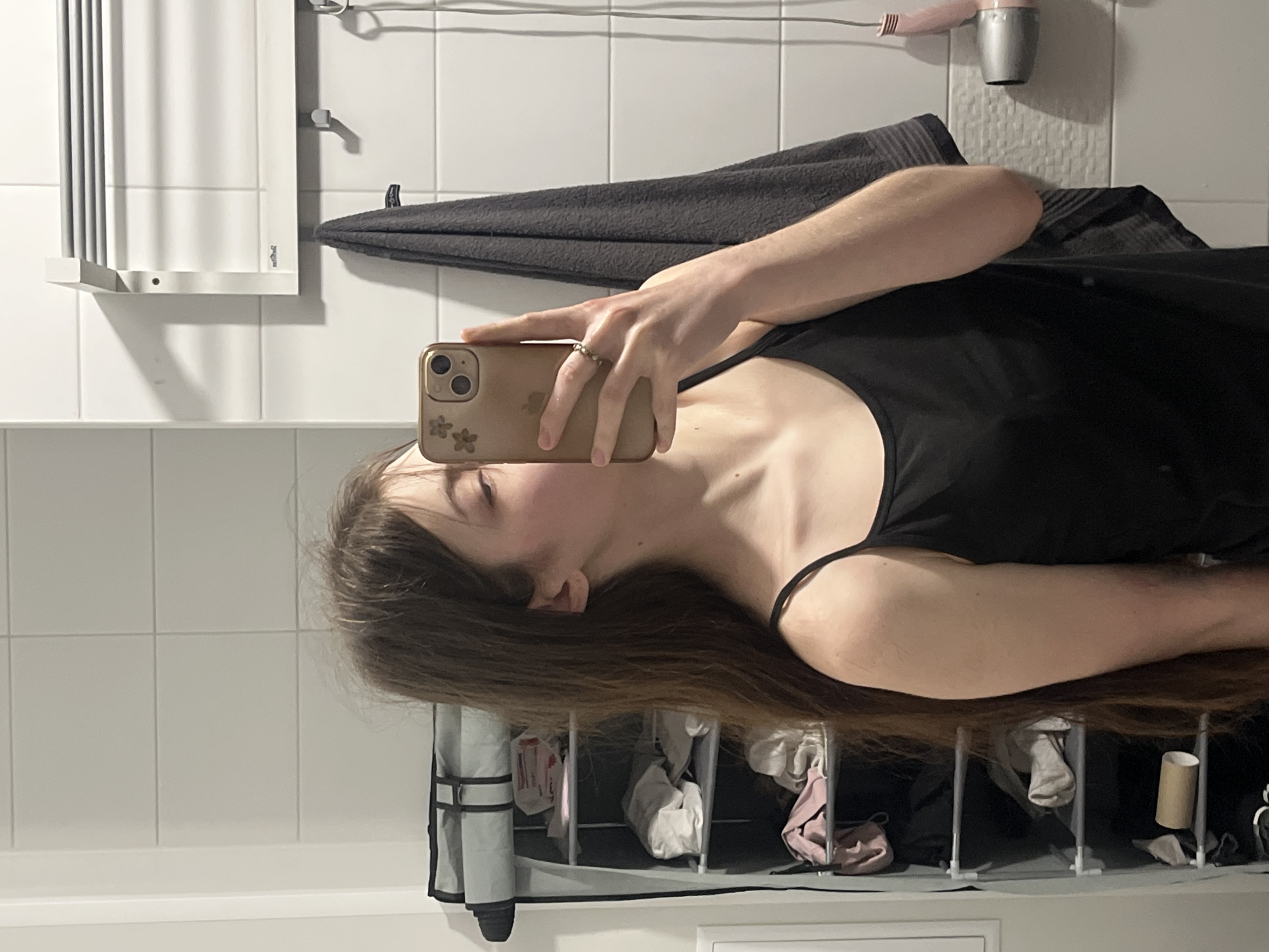 A person with long brown hair is holding a mobile phone, presumably taking a selfie in a bathroom. The bathroom has white tiled walls and a heated towel rack. A gray towel hangs on a towel rack. A hair dryer is visible on the wall. Below the person, a storage unit is visible, containing various objects including a cardboard tube. The image provides no information about the location of the city of Saint Petersburg, Russia.