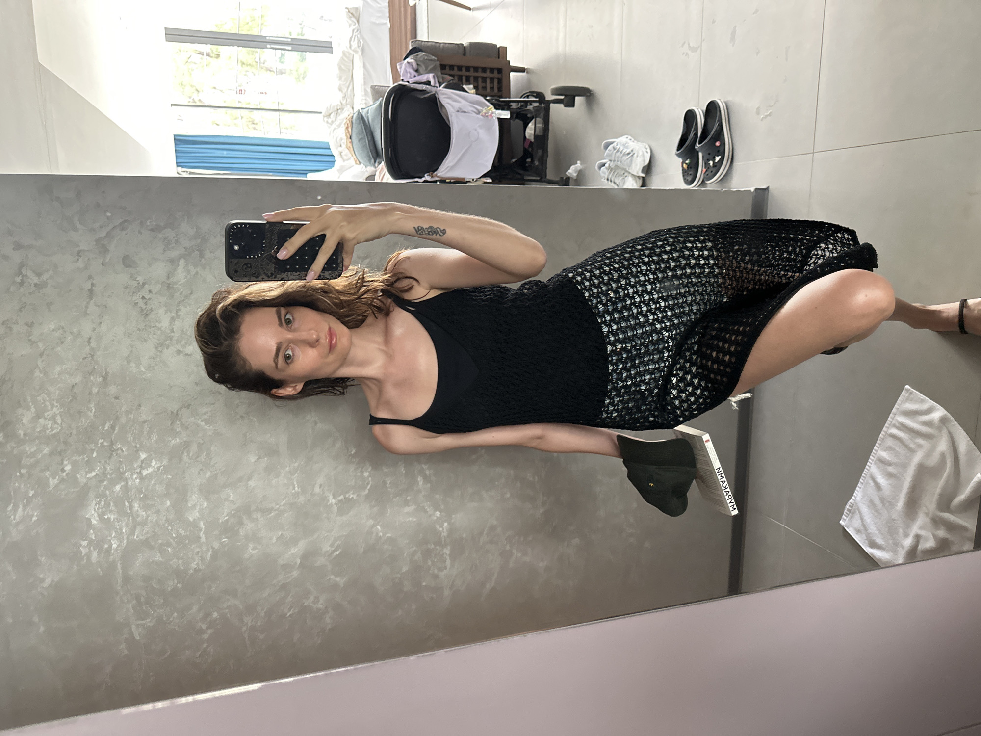 A woman is taking a selfie in a bathroom mirror in Choeng Thale, Thailand. She's holding a phone with a patterned case in her left hand. She has brown hair and is wearing a black top and a black mesh skirt. Reflected in the mirror are a sink with gray textured walls, a towel, a book, and a green hat. In the background, through a window, are glimpses of a building exterior, possibly a residential structure, and what appears to be baby furniture and scattered objects. Shoes are visible on a ledge or wall. The lighting appears to be bright, natural daylight. The bathroom is somewhat cluttered.
