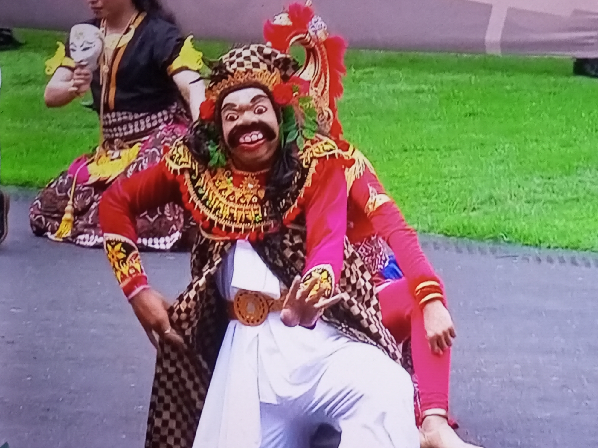 The image shows two individuals in traditional attire, likely performers, on a paved surface in front of a grassy area. The primary subject is in the foreground, a person wearing an elaborate headdress and a mask with a wide, exaggerated smile, dark mustache, and large eyes. This performer is dressed in a red top with decorative gold trim and a patterned sarong or skirt that includes black and white checks and colored fabrics. A white garment is worn underneath. The performer's left arm is bent at the elbow, and their hand is positioned near their chest, adorned with a gold circular ornament. A brown, circular belt with a woven pattern is visible around their waist.
To the left of the main performer, a second individual is partially visible, also in traditional clothing. This person wears a black top with yellow accents on the shoulders and holds a white mask in their left hand. A colorful, patterned skirt is also visible. The background consists of a neatly trimmed green lawn. The overall scene suggests a cultural performance or ceremony taking place outdoors. The location is specified as Banyudono, Indonesia.