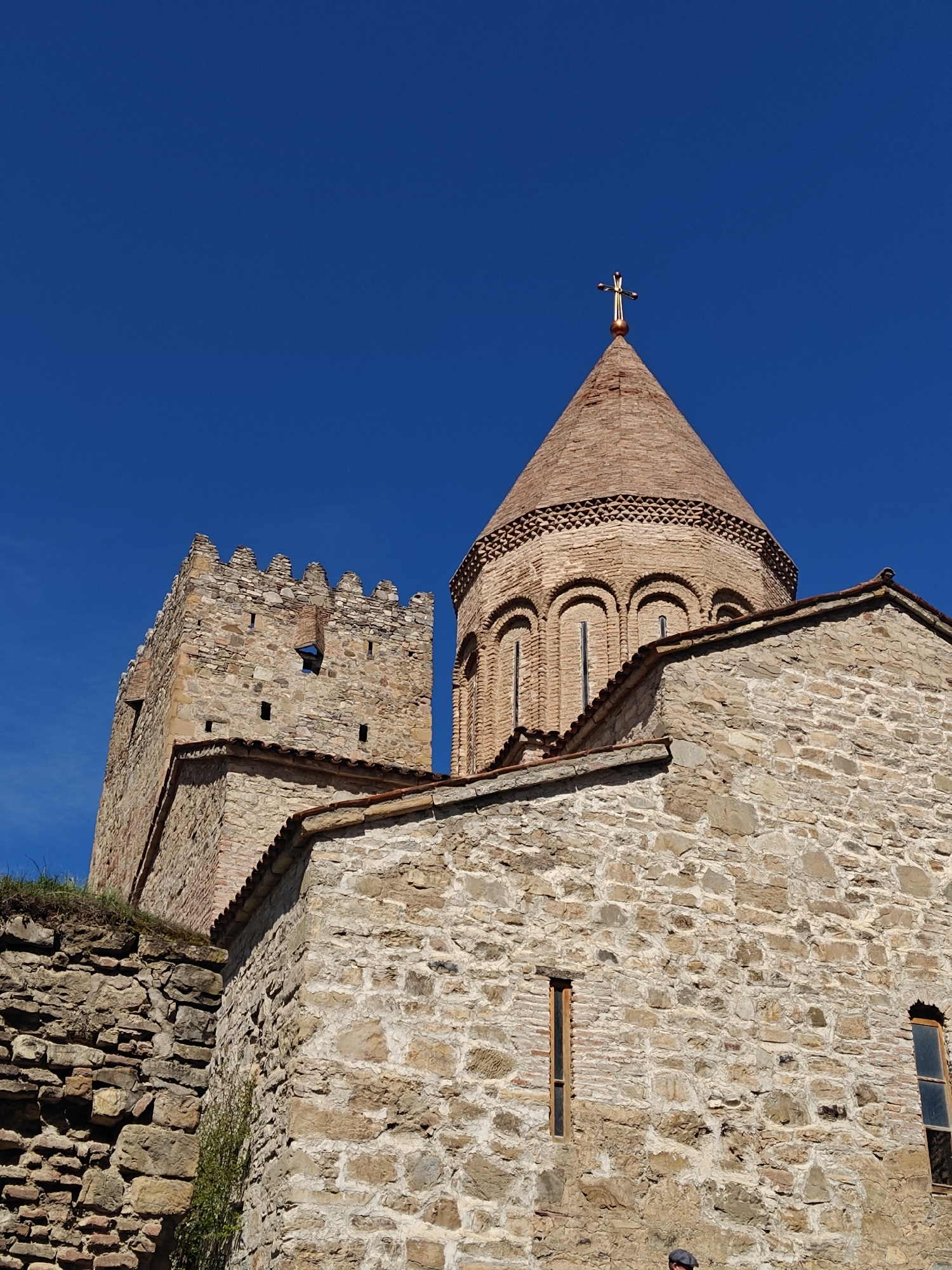 The image shows the Ananuri fortress, located in Georgia. The architectural complex consists of two churches, a tower, and defensive walls. The churches and the tower are constructed from stone. The walls show different kinds of construction. The sky is clear and blue. The picture is taken from below.