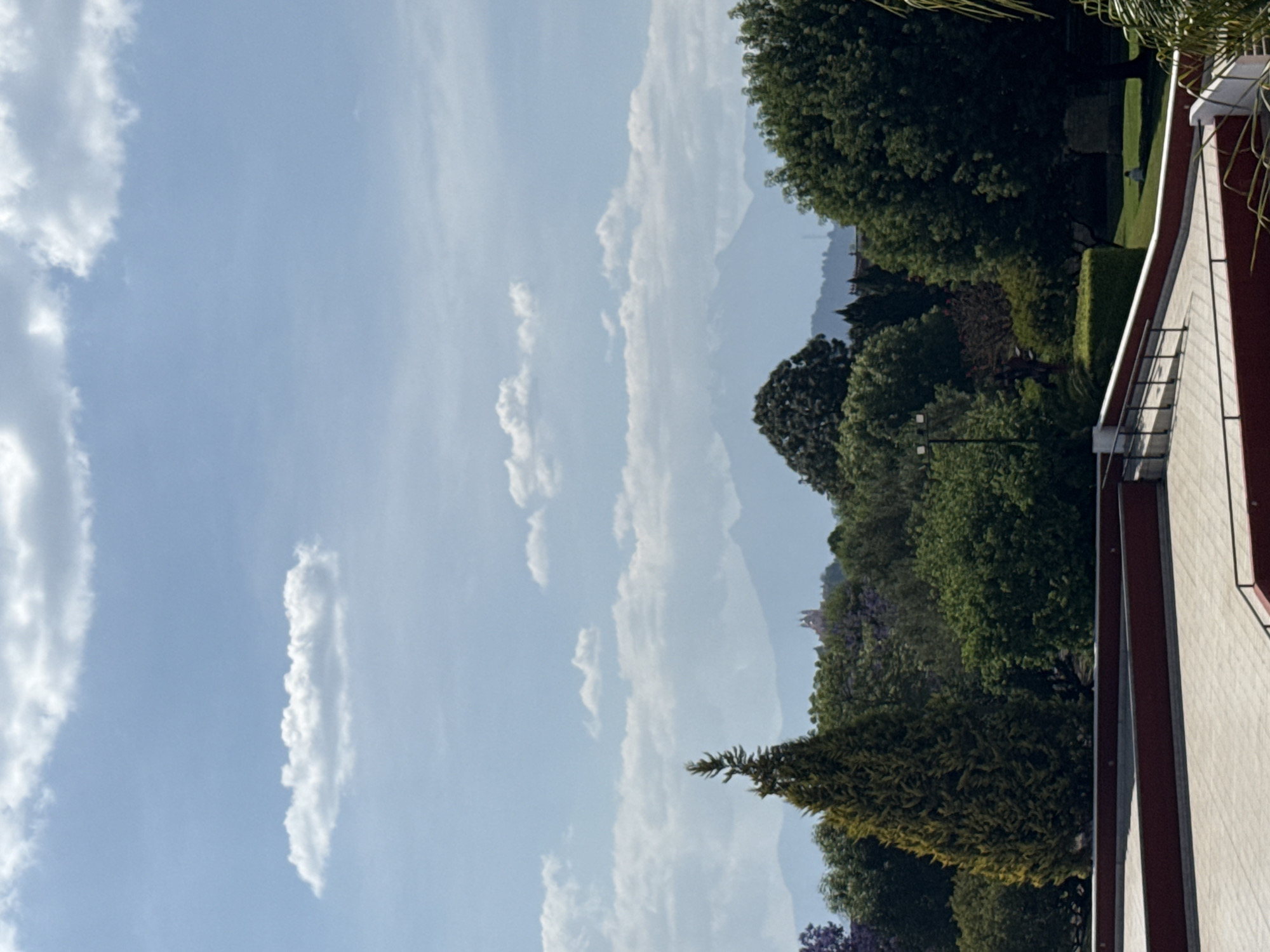 The image shows a landscape in San Andrés Cholula, Mexico. In the foreground is a building with a red-trimmed white wall and a metal railing. The background reveals a lush green landscape filled with trees of various shapes and sizes. There is a view of mountains in the distance, shrouded in a haze. Above, a bright blue sky is scattered with elongated white clouds. The photo has been rotated 90 degrees.