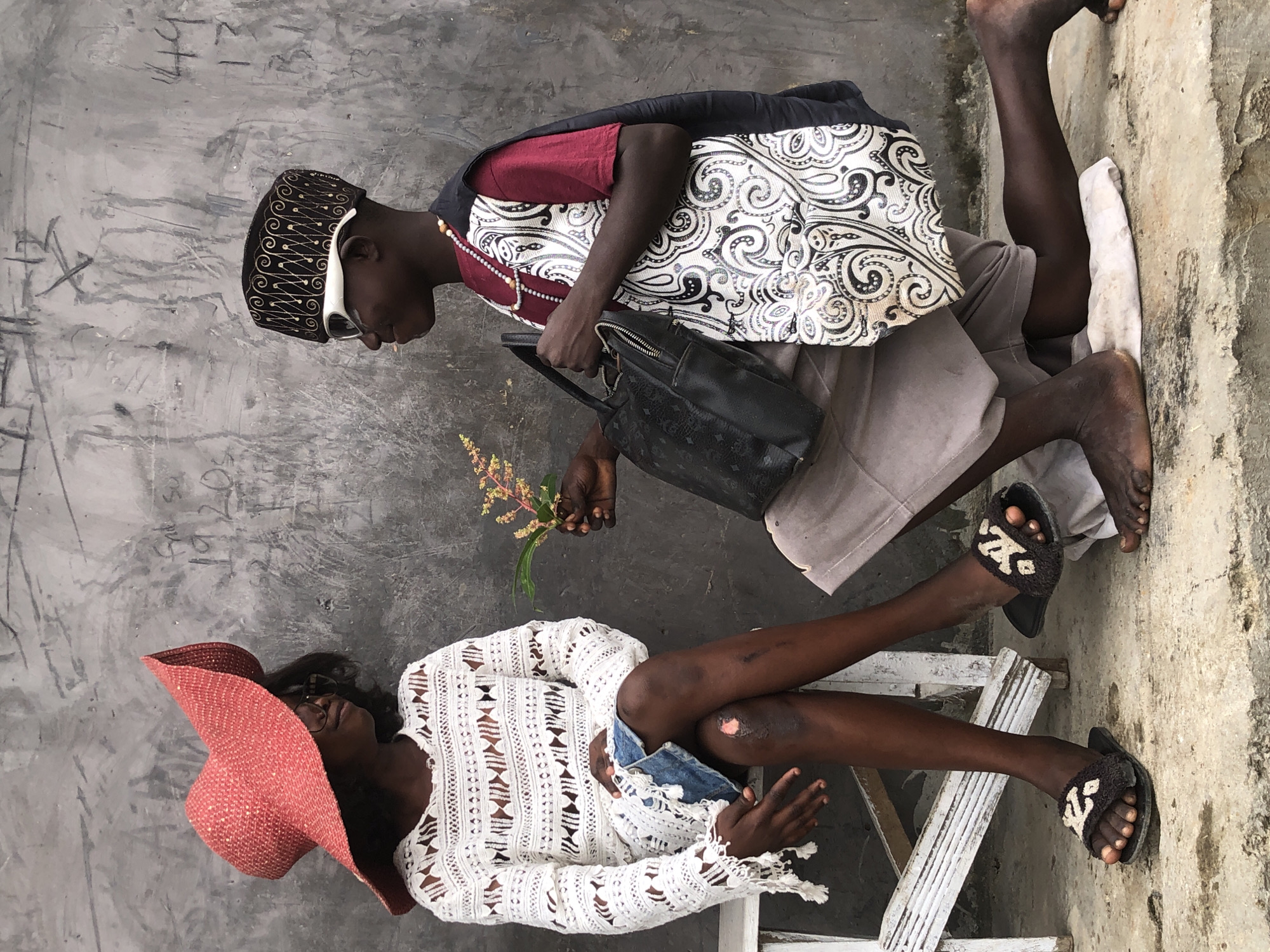 The image depicts a young boy offering a sprig of flowers to a young girl. The scene is set against a rough, grey concrete wall with faint markings. The girl, seated on a white wooden bench, wears a large, wide-brimmed red hat and a white, open-knit long-sleeved top over a denim skirt. Her legs are crossed, and a scraped knee is visible. She wears dark, open-toed sandals with a decorative pattern. The boy, kneeling on a white cloth, is dressed in a maroon t-shirt, a decorative black and white vest, and a patterned cap. He wears white-rimmed glasses and a beaded necklace. He holds a black handbag in his left hand and offers the flowers with his right. The boy's bare feet are visible. The ground is concrete, and there are some minor stains. The overall impression is a staged scene with a tender gesture. The image is located in Ado, Nigeria.
