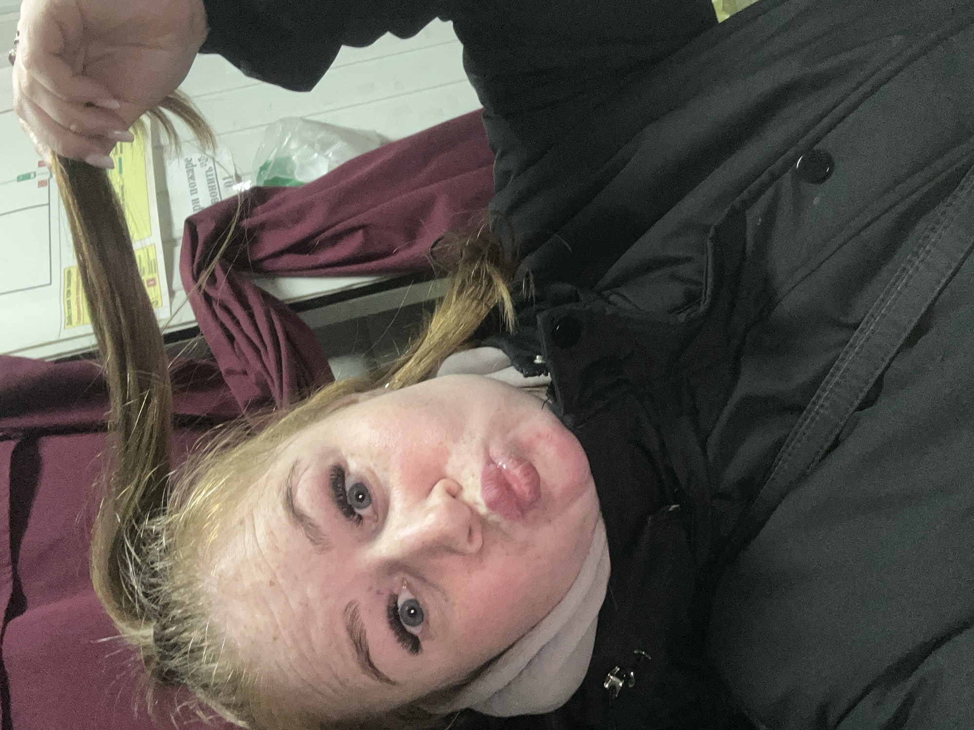 A woman with blonde hair is holding a strand of her hair up in her hand. She is wearing a black jacket and a white scarf. She is lying on a bed with maroon sheets. There is a white wall in the background with a sign on it. The location is Chaikovsky, Russia.