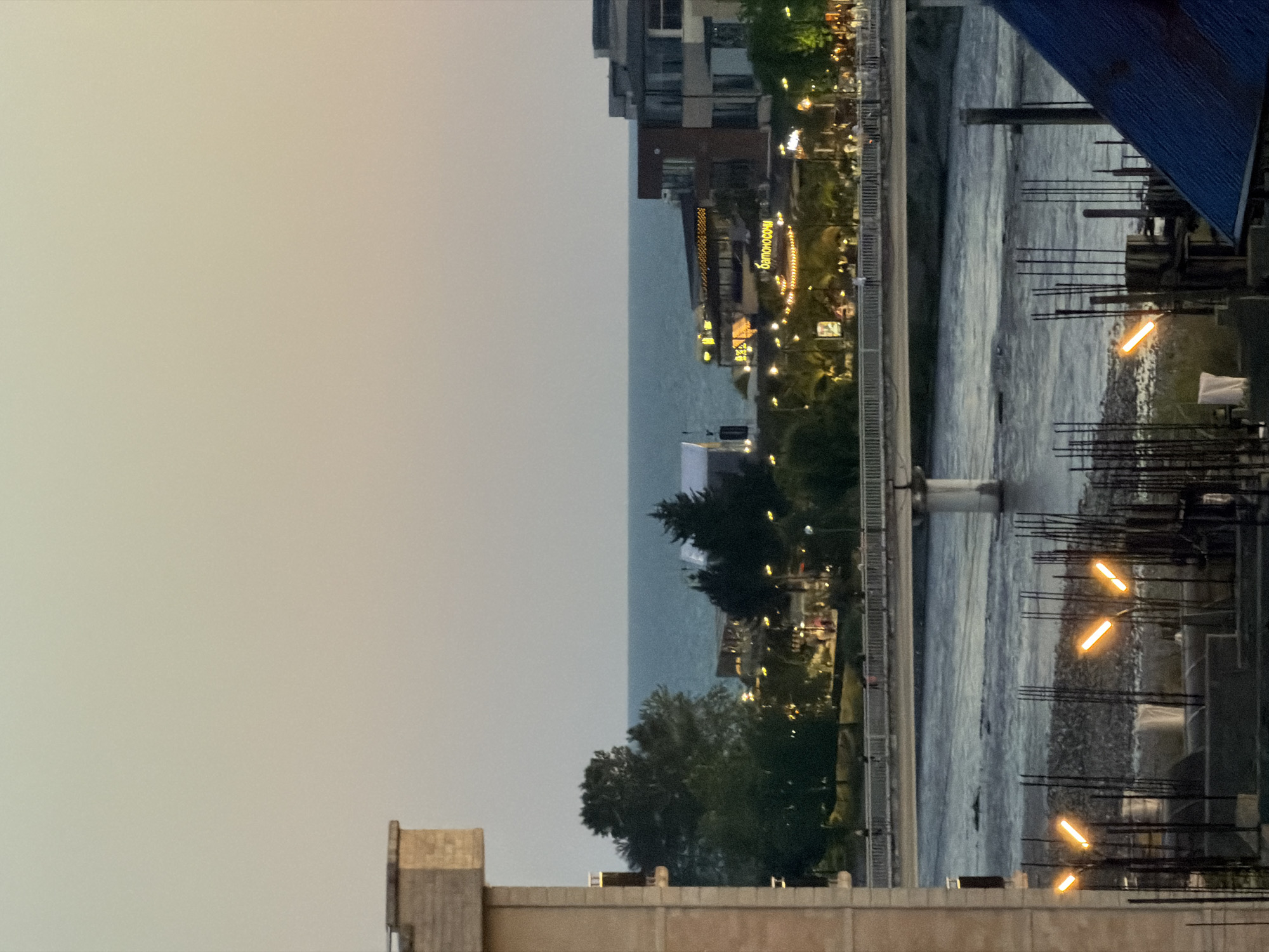 An elevated shot captures a waterfront cityscape in Sirius, Russia, at dusk. In the foreground, construction scaffolding with rebar and some plastic sheeting is visible, with several orange construction lights illuminating the area. Beyond this, a wide river flows towards the sea, crossed by a pedestrian bridge with lights. On the opposite bank, a promenade is lined with trees, shops, and restaurants, their lights twinkling in the fading light. The sea stretches out in the background, meeting a hazy sky. The overall impression is of a developing urban area by the coast transitioning into evening.
