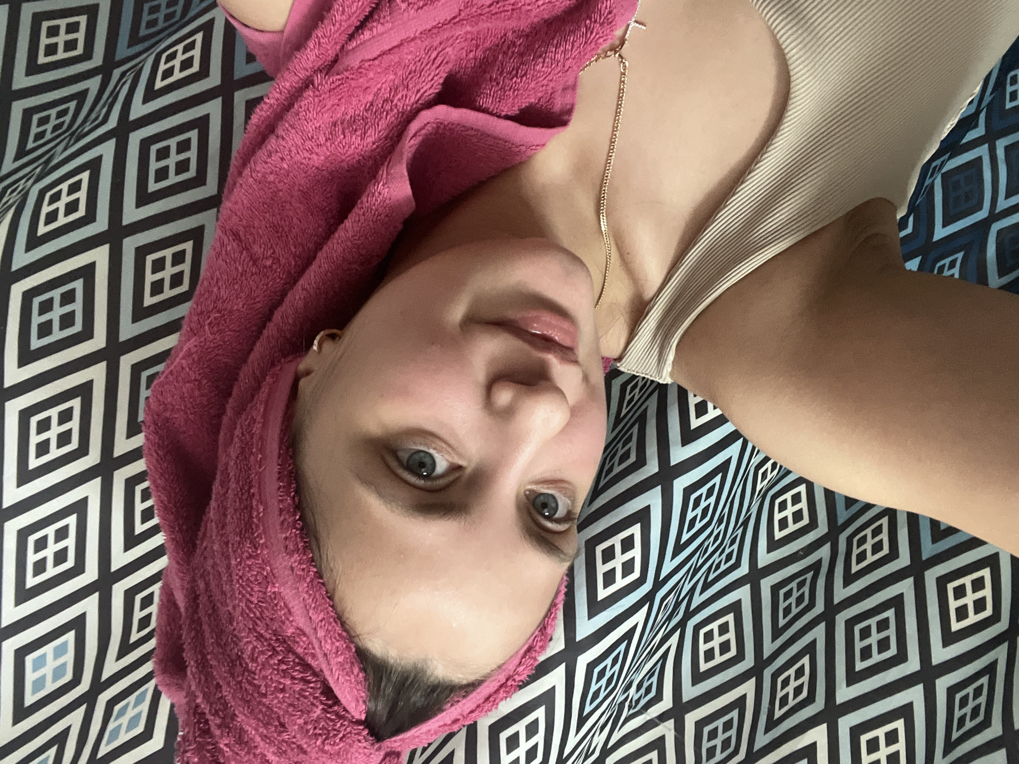 The media file shows a close-up, sideways, overhead shot of a young woman lying down. She has a pink towel wrapped around her head, and her eyes are open, looking towards the camera. Her lips are slightly parted, and she has a light dusting of makeup on her face, including lip gloss. She is wearing a ribbed, cream-colored tank top. A thin gold chain necklace is visible around her neck. The background is a patterned fabric with diamond shapes in shades of blue, black, and white. The overall impression is of a person relaxing or preparing for a spa treatment. The location context provided is Марковский, Russia. There is no visible text in the image.