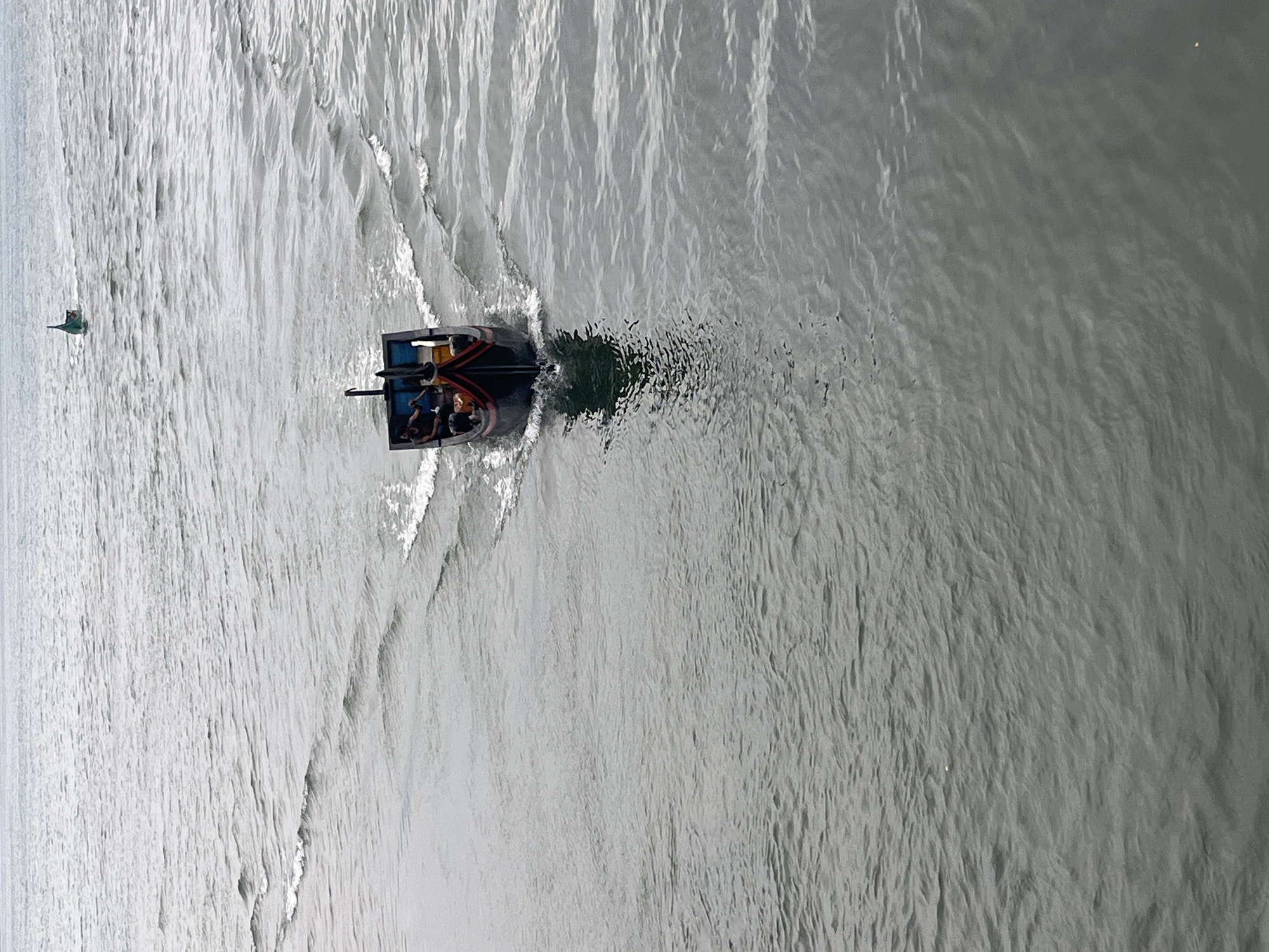 The image captures a calm, overcast day at sea. In the foreground, a dark-colored boat with red trim navigates through gentle waves, leaving a visible wake. Several people are seated inside the boat. In the background, further out at sea, another, smaller green boat is visible. The water appears grey and rippled. The scene is likely a coastal area of Banda Aceh, Indonesia, given the general appearance of the water and the type of boat.