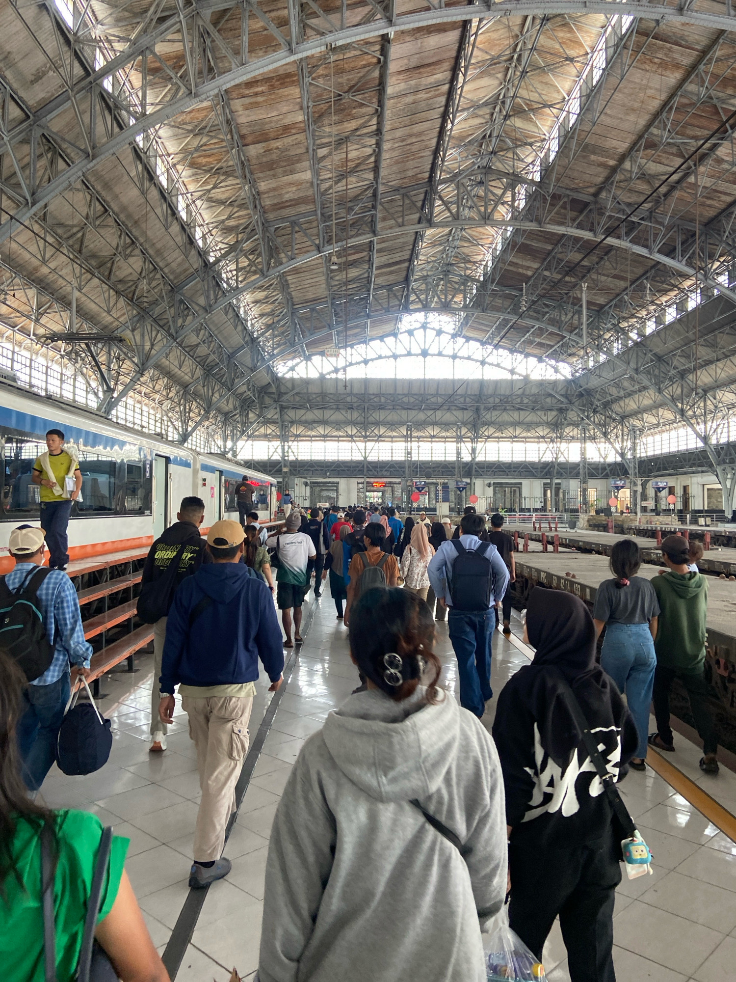 The image captures a bustling scene inside a train station, likely in North Jakarta, Indonesia, characterized by its expansive, industrial-style arched roof supported by a complex network of metal trusses and beams. Sunlight streams through the translucent panels of the roof, illuminating the vast space.
A train, predominantly white with a blue and orange stripe along its lower half, is visible on the left side of the frame, parked at a platform. Several people are disembarking from or waiting near the train.
A significant number of people are walking in both directions along the platforms, many carrying bags or backpacks, suggesting a busy travel day. The floor is tiled in a light color with dark lines marking pathways. The overall atmosphere appears to be one of movement and transit. The architecture suggests a large, established train station.