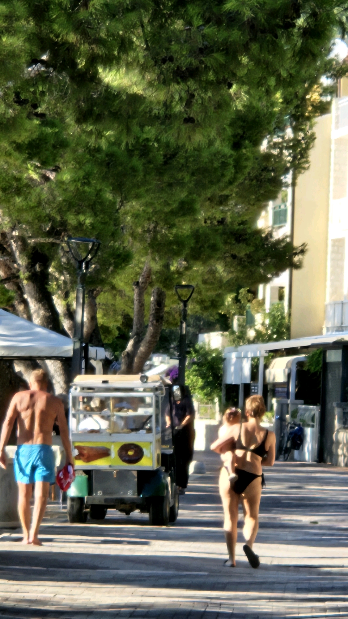 In Općina Tučepi, Croatia, on a sunlit, paved walkway lined with trees and buildings, two individuals are walking. In the foreground, a man with a muscular physique, wearing blue swim trunks, walks away from the camera, holding a red object. To his left is a mobile food cart, appearing to sell donuts and other items. Further down the walkway, a woman in a bikini walks away from the camera, carrying a child. The woman is wearing a black bikini top and black bikini bottoms, and the child is being held in her arms. A black umbrella or object is visible on the food cart. The environment suggests a tourist area, possibly a promenade or street near a beach.