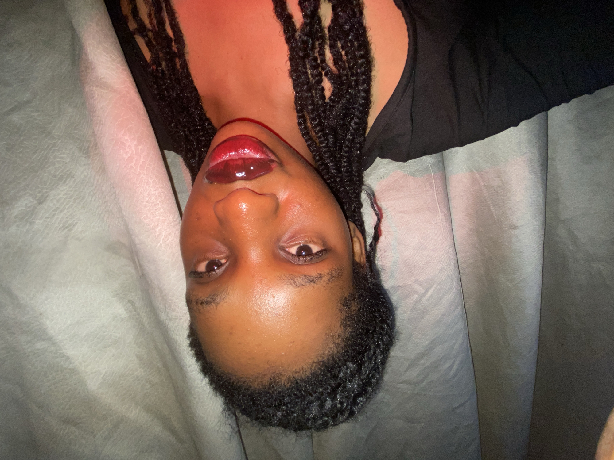 A close-up, upside-down, medium shot depicts a person with dark skin and dark, braided hair, wearing a dark top, lying on a textured, light-colored fabric. The person has glossy, red lips and their gaze is directed towards the camera. The location and city are unknown.