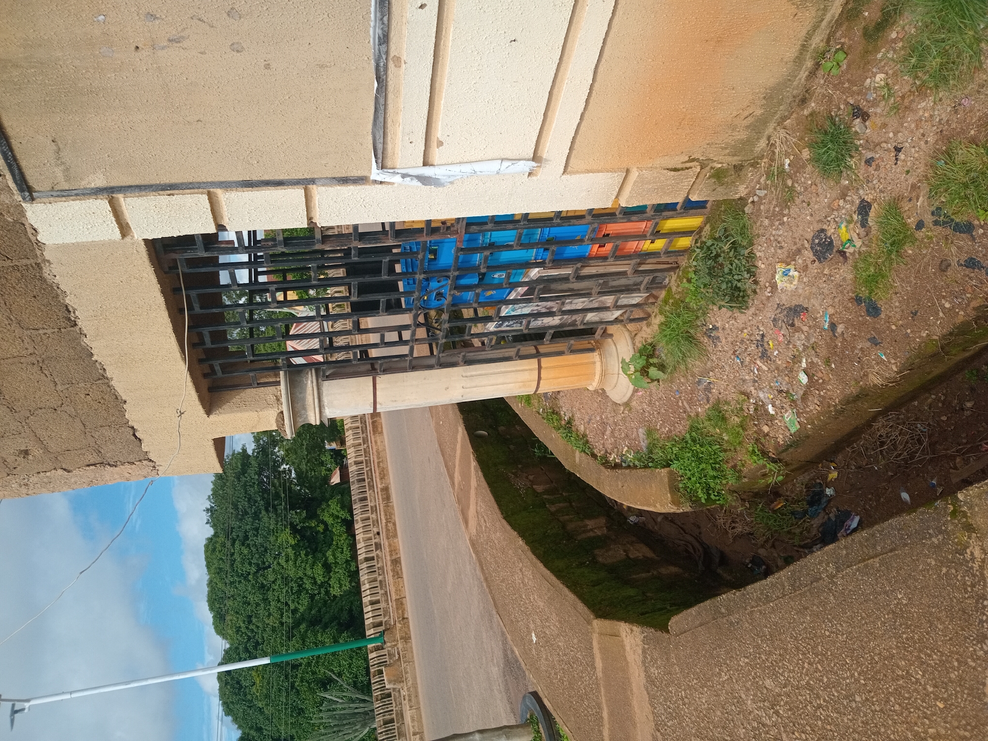 The image depicts an outdoor scene in an urban environment, likely in a city located in Unknown, Unknown. In the foreground, a concrete drainage ditch runs along the side of a road, filled with debris and some overgrown vegetation. To the right of the ditch is a building with a textured, light brown facade. A window opening in the building is covered by a black metal grate, behind which several colorful plastic crates (blue, yellow, and orange) are stacked. A white cloth is draped over the top edge of the building's facade to the right of the grate. Further in the background, a road curves past, with a stone barrier and lush green trees lining the other side. A tall white pole with a green section at the bottom stands near the road, and some large, spiky plants are visible to its left. The sky is partly cloudy, with patches of blue visible. There are no people visible in the image.