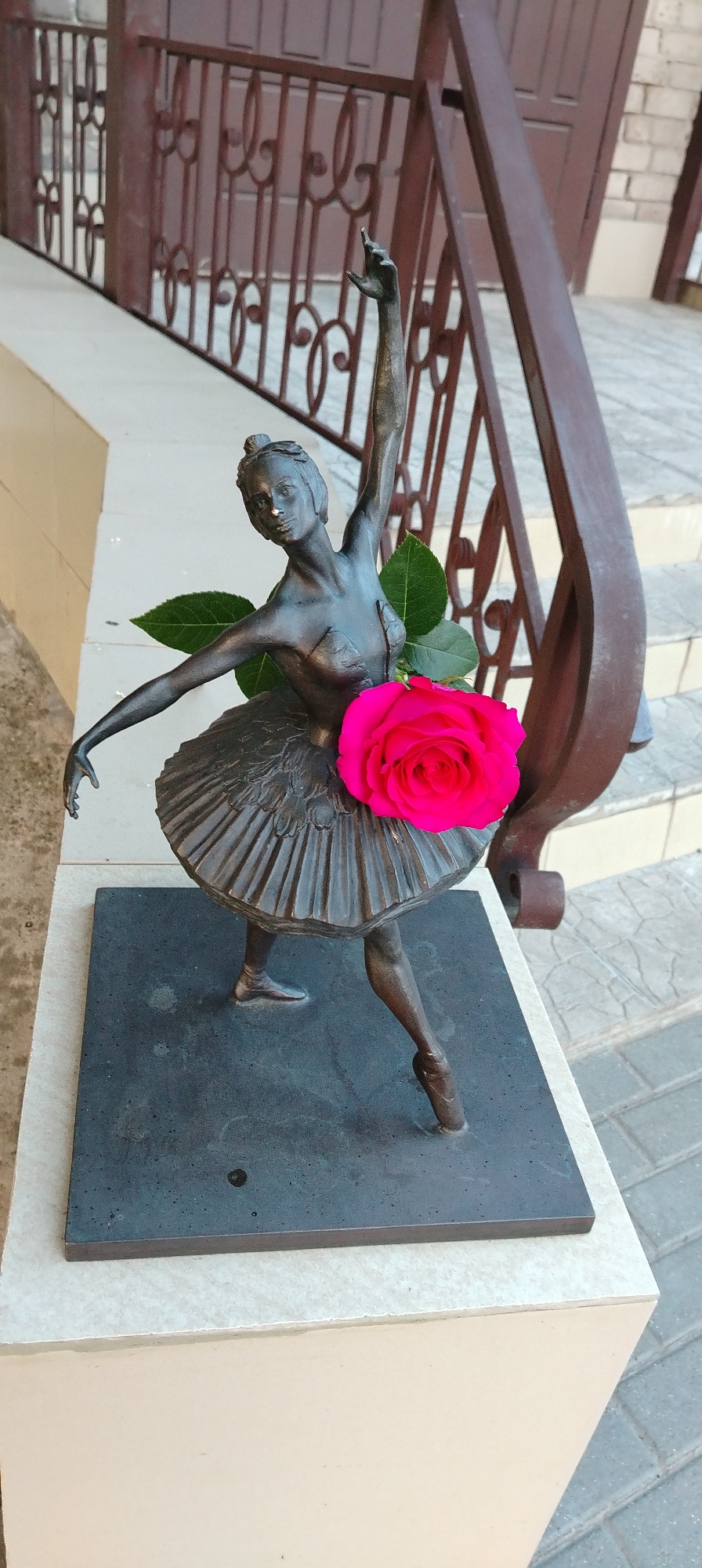A close-up, slightly high-angle shot shows a bronze statue of a ballerina with a bright pink rose adorning her tutu. Two green leaves frame the rose. The statue is placed on a dark square base that sits atop a cream-colored ledge. Behind the statue, a dark brown metal railing with intricate loops and swirls is visible, and steps are partially visible behind the railing. The setting is outdoors, with brick pavers and a light-colored wall in the background. The photograph depicts the ballerina statue located in the city of Chaikovsky, Russia.