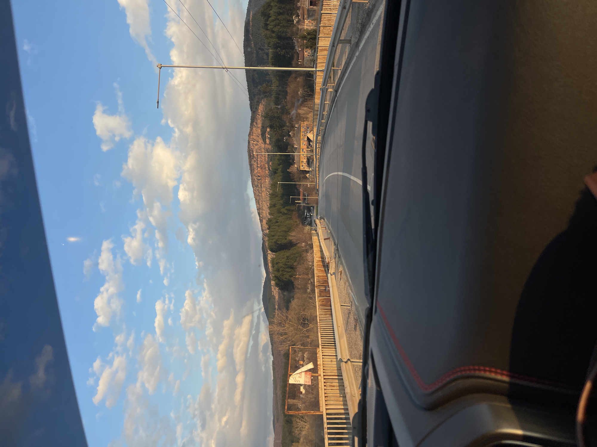 The image shows a view from inside a vehicle, likely a car, driving on a bridge or elevated road in Batanovtsi, Bulgaria. The road curves slightly to the right. A white dividing line is visible on the asphalt. Outside the vehicle, there's a mountain with sparse vegetation. Overhead power lines run parallel to the road. A metal guardrail separates the road from the adjacent terrain. The sky is visible with scattered clouds. A sign is seen, though unreadable due to distance and resolution. The vehicle's dashboard and windshield are visible in the foreground.