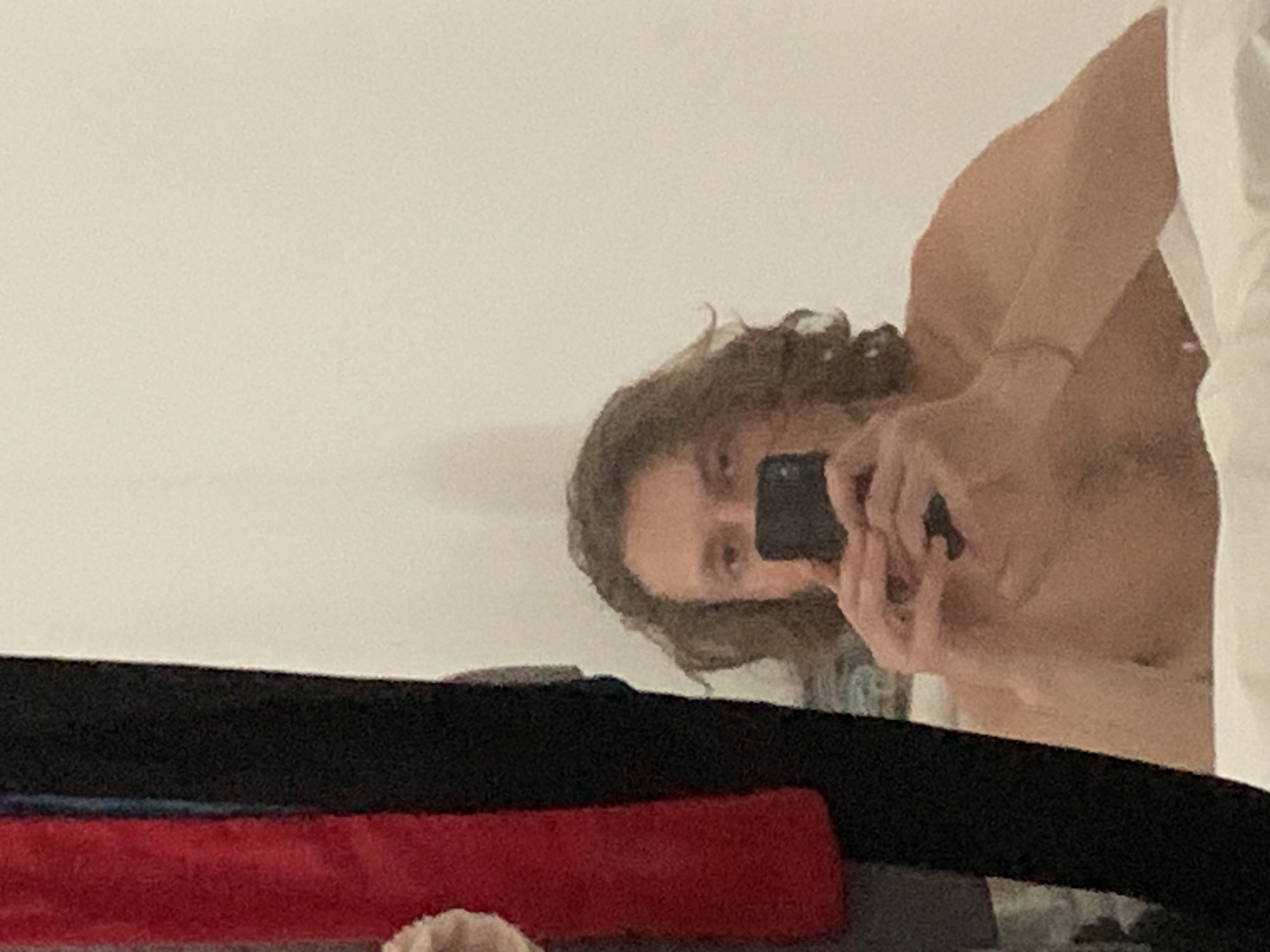 The image displays a mirror selfie of an individual in an indoor setting. The specific city location of Tel-Aviv, Israel, as requested, cannot be confirmed from the visual information provided within the image.
**People:** One individual, appearing male, with medium-length, wavy brown hair, is visible from the upper chest upwards. The individual's face is largely obscured by a smartphone held directly in front of it; only the eyes and forehead are partially visible above the device. The individual appears to be shirtless.
**Subjects/Objects:** The central subject is the individual's reflection. The individual holds a black smartphone horizontally with both hands, positioned to capture the image. A thin, light-colored bracelet or band is present on the individual's left wrist. In the foreground, below the individual's torso, white bedding or sheets are visible, suggesting a bed or similar soft surface. To the left of the reflected individual, hanging or draped textiles are visible: a prominent section of textured red fabric is next to a solid black fabric, with other indistinct darker fabrics possibly behind them. The background behind the individual's reflection consists of a plain, light beige or off-white wall.
**Actions/Interactions:** The individual is actively taking a picture of their reflection in a mirror using the smartphone. Both hands are engaged in holding the device.
**Environment:** The scene takes place indoors, likely a private room such as a bedroom or hotel room, inferred from the presence of bedding and a plain wall. Lighting appears even, suggesting an interior light source.