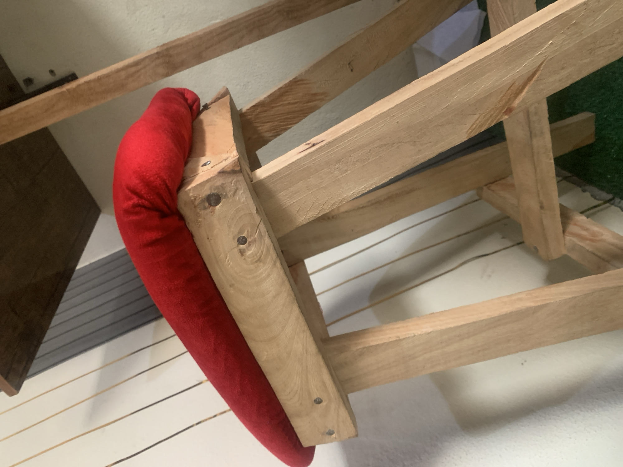 A close-up, angled shot shows a red bolster pillow resting on a wooden easel. The pillow is cylindrical and draped over the horizontal support of the easel. The easel is constructed from light-colored wood and appears to be handmade, with visible screws and rough edges. The background consists of a textured white wall and a dark wooden shelf above. Vertical lines, possibly cords or thin wooden planks, are visible against the wall behind the easel. The lighting is soft, casting subtle shadows. The image does not depict any specific urban or natural environment within Jauro Yinu, Nigeria, as it focuses solely on the object and its immediate surroundings.
