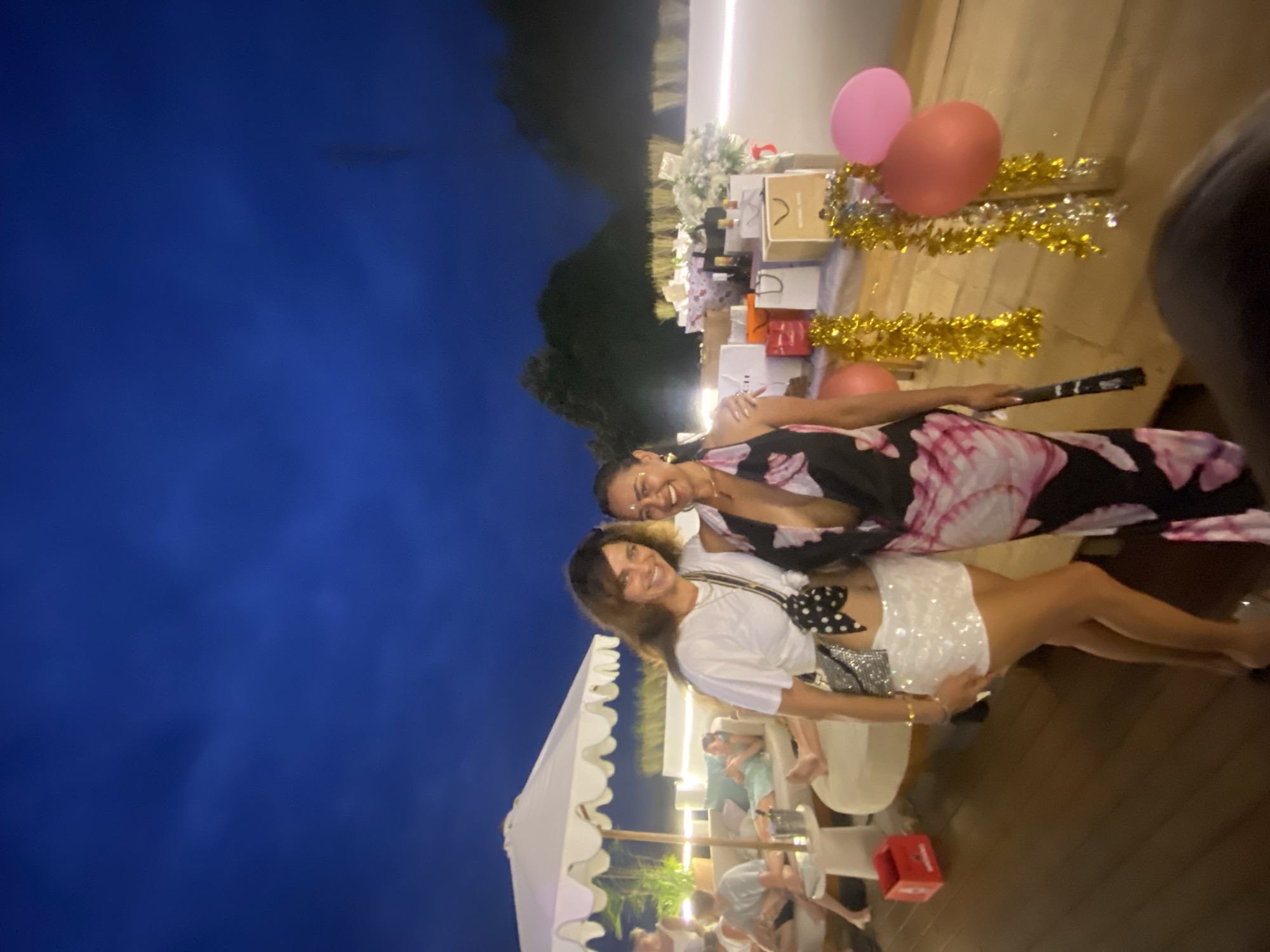 This image depicts a nighttime outdoor gathering on a wooden deck in Nusa Penida, Indonesia. Two women, the primary subjects, are posing for the camera. The woman on the left wears a white t-shirt, a sequined white skirt, and a black and white polka dot bag. The woman on the right is dressed in a black and pink floral dress with a deep V-neck. Behind them, several gift bags are arranged on a table, adorned with gold and silver tinsel. In the background, there are other people relaxing on lounge furniture under a white umbrella. The dark blue night sky with faint clouds forms the backdrop, and distant hills are visible. Two balloons, one pink and one red, are situated near the gift table. The overall atmosphere suggests a celebration or party.